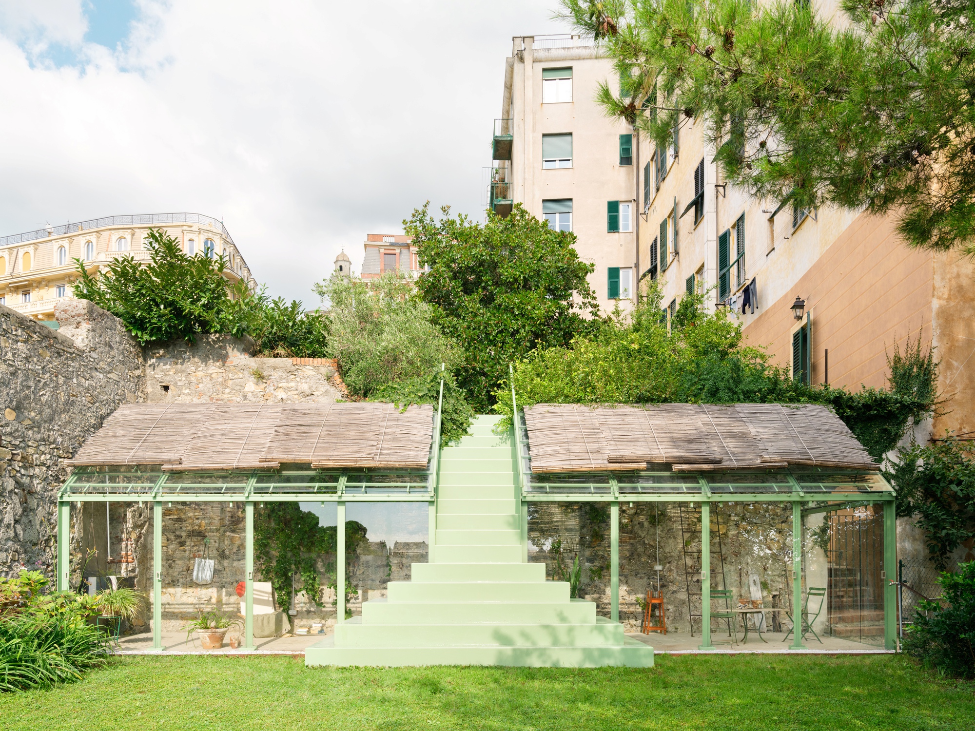 Front view of two glazed green-framed pavilions with reed roofs, split by a pale green central staircase, set against a stone wall and apartment blocks