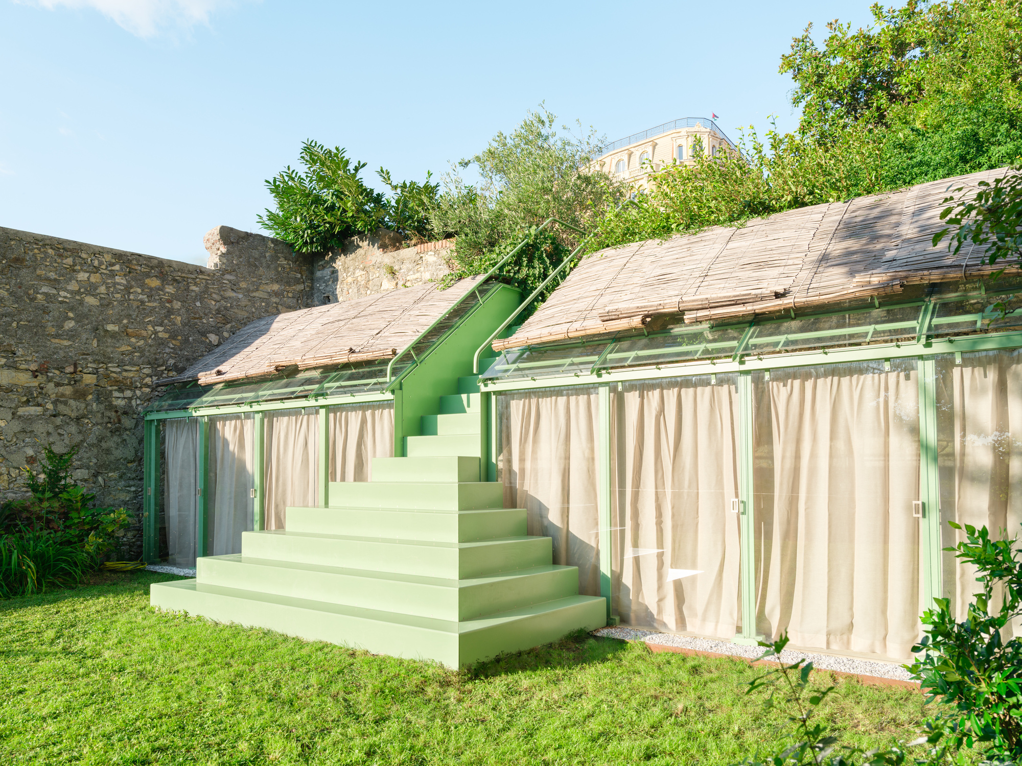 Side view of the green stepped staircase between reed-roofed glass pavilions with linen curtains, beside an old stone wall in bright afternoon sun