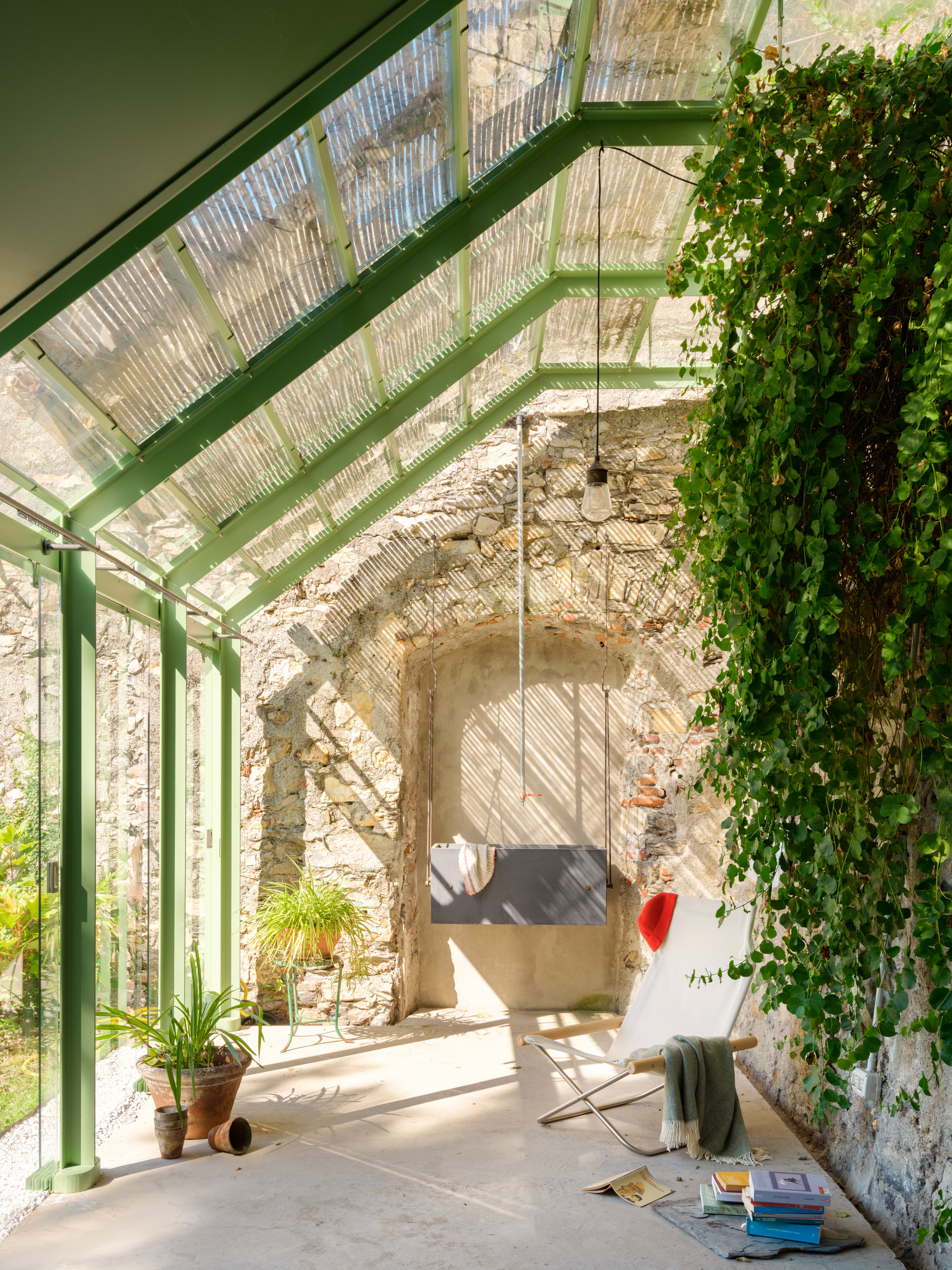 Interior of the glazed room with a folding lounge chair, hanging lamp and trailing ivy against a vaulted stone niche in raking sunlight