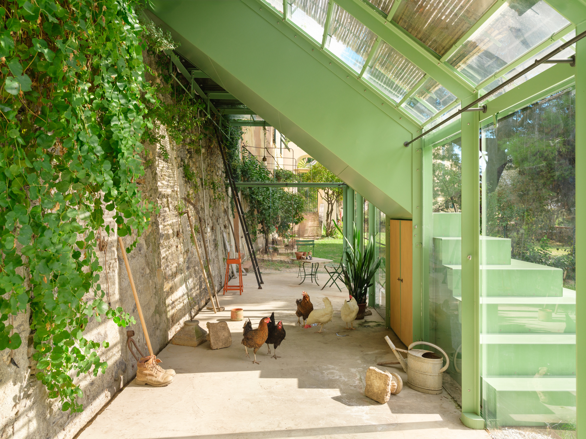 Concrete-floored interior beneath the green staircase, with hens, a watering can, a ladder and ivy hanging over the stone wall