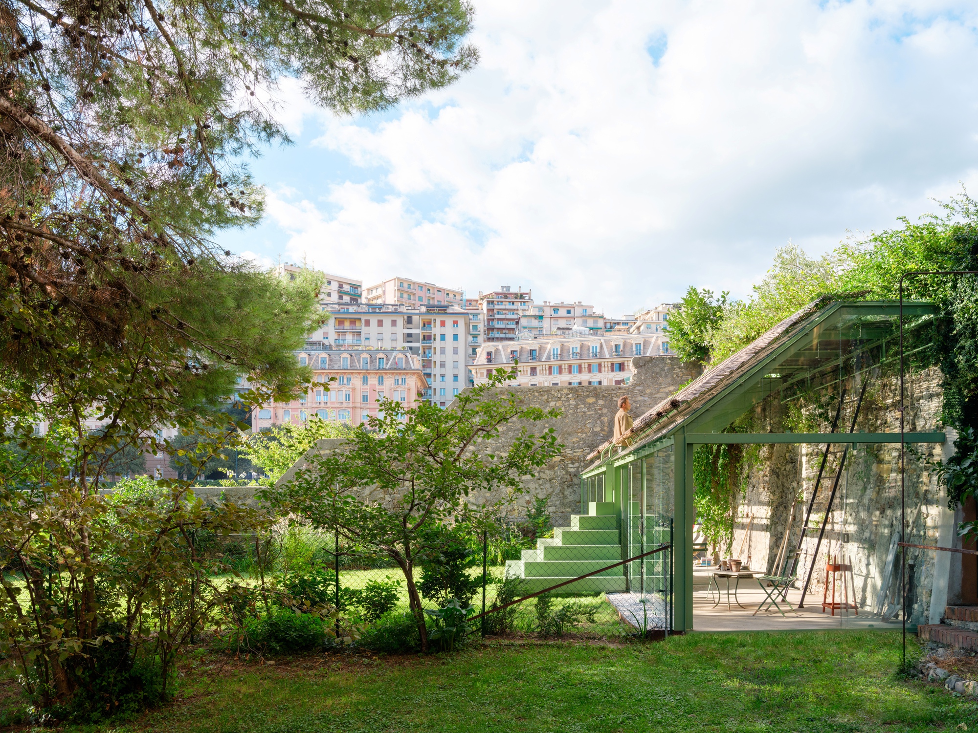 Green steel-framed glass pavilion with a reed-covered roof against a stone wall, seen across a lawn with apartment blocks behind