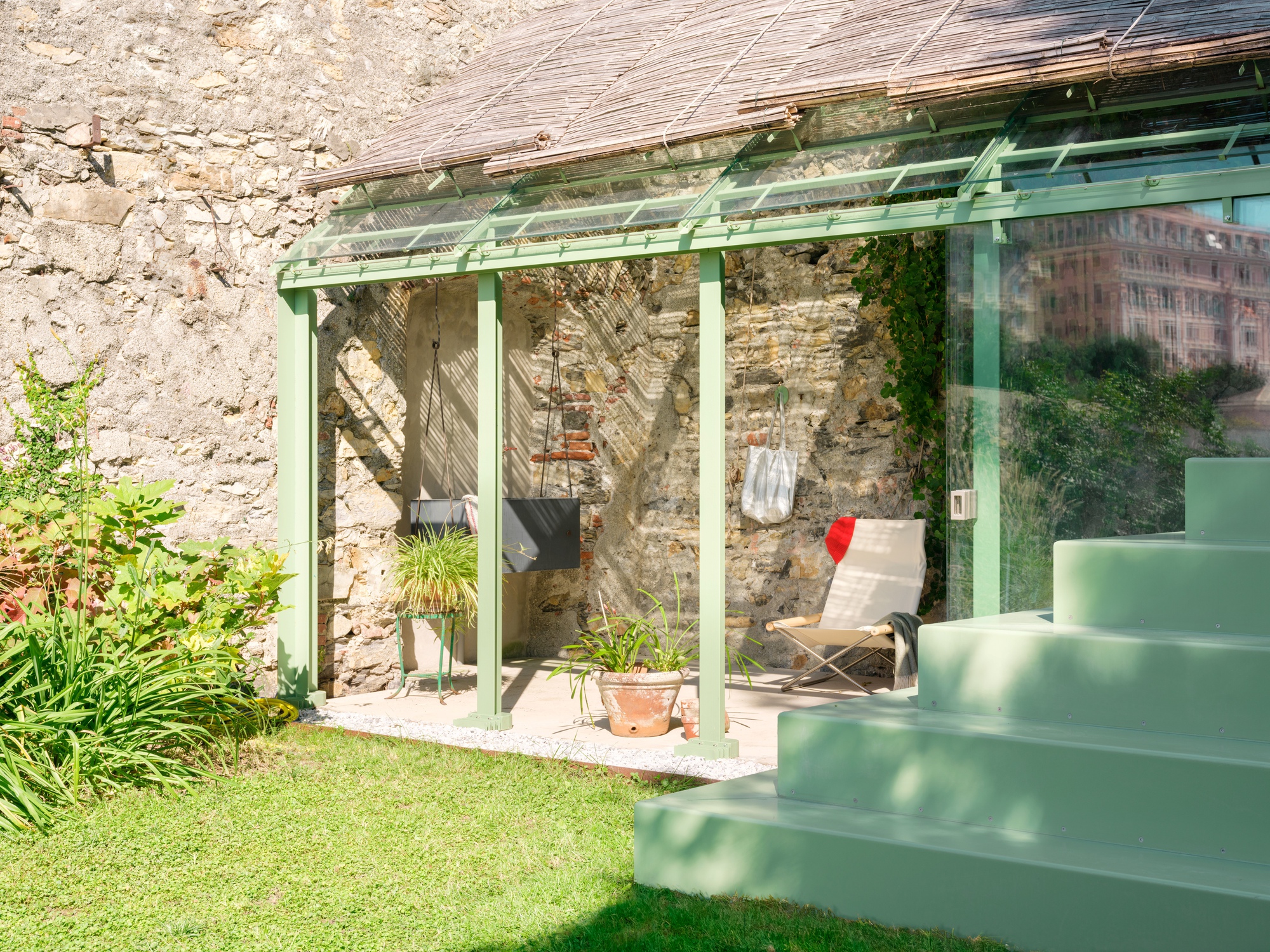 Open side of the pavilion showing a lounge chair, potted plants and a hanging seat against a rough stone wall in sunlight
