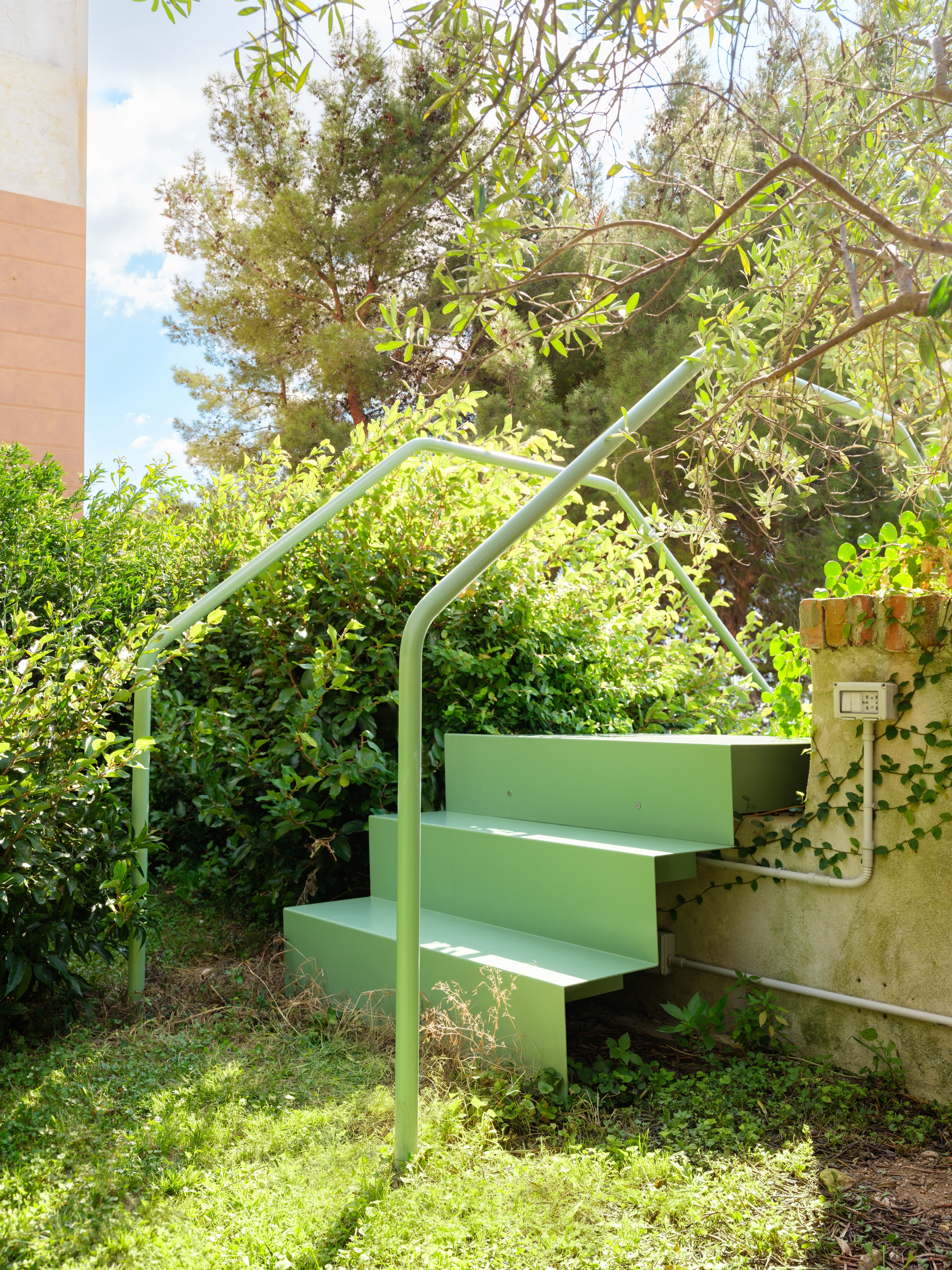 Three-step green metal stair with looped handrails beside a plastered garden wall, shaded by an olive branch and pine