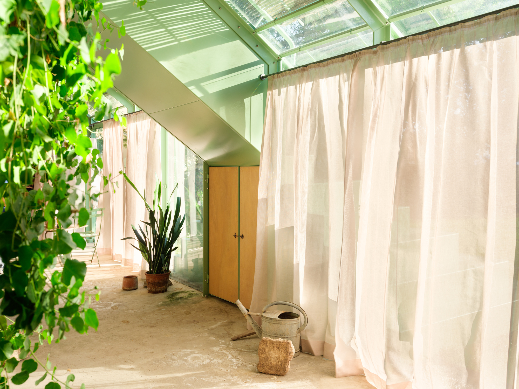 Sheer white curtains drawn along the glazed wall beside a timber cabinet, a watering can and a potted plant in warm light