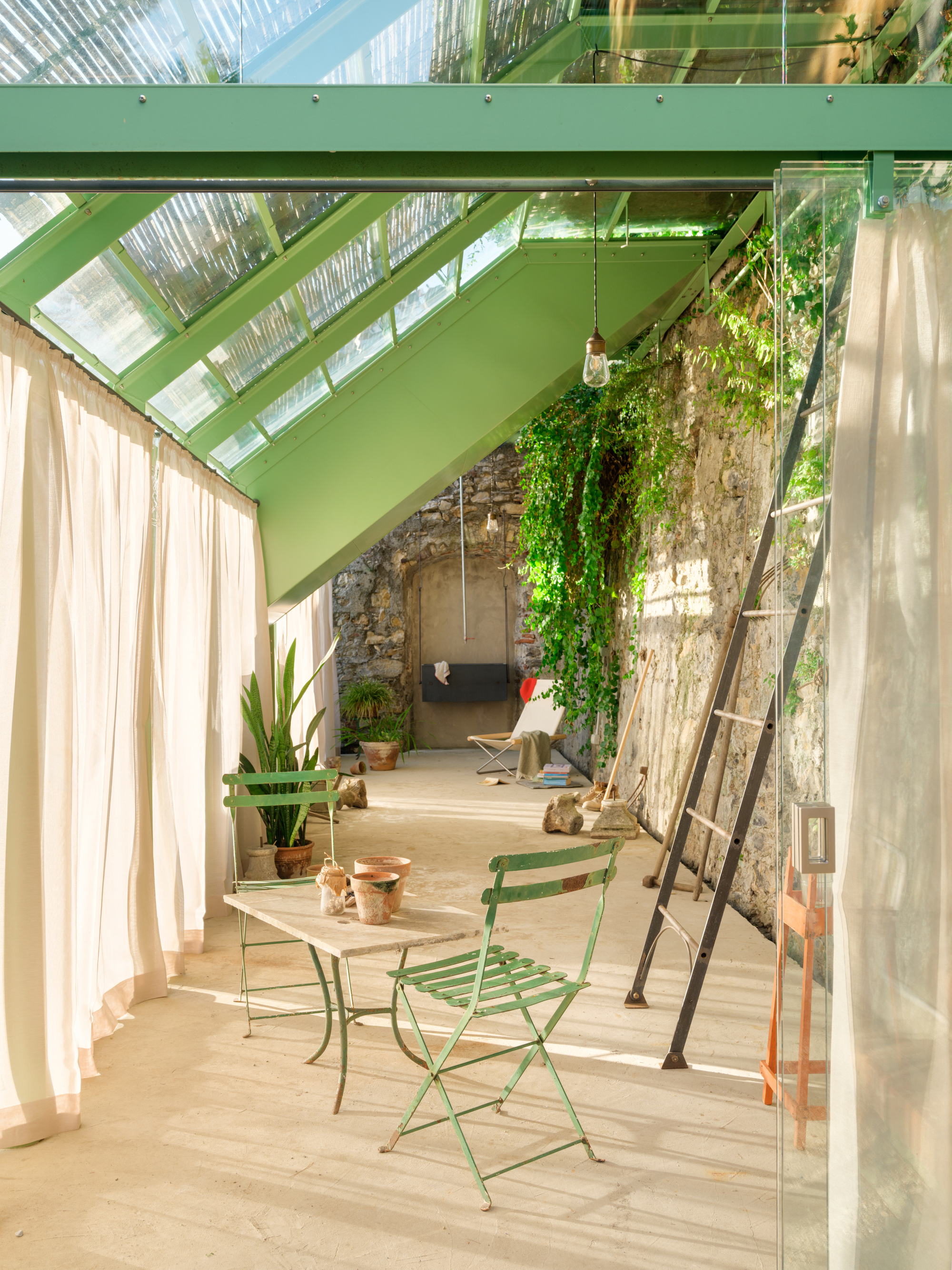 Two folding green metal chairs and a small table under the glass roof, with a leaning ladder and ivy on the stone wall
