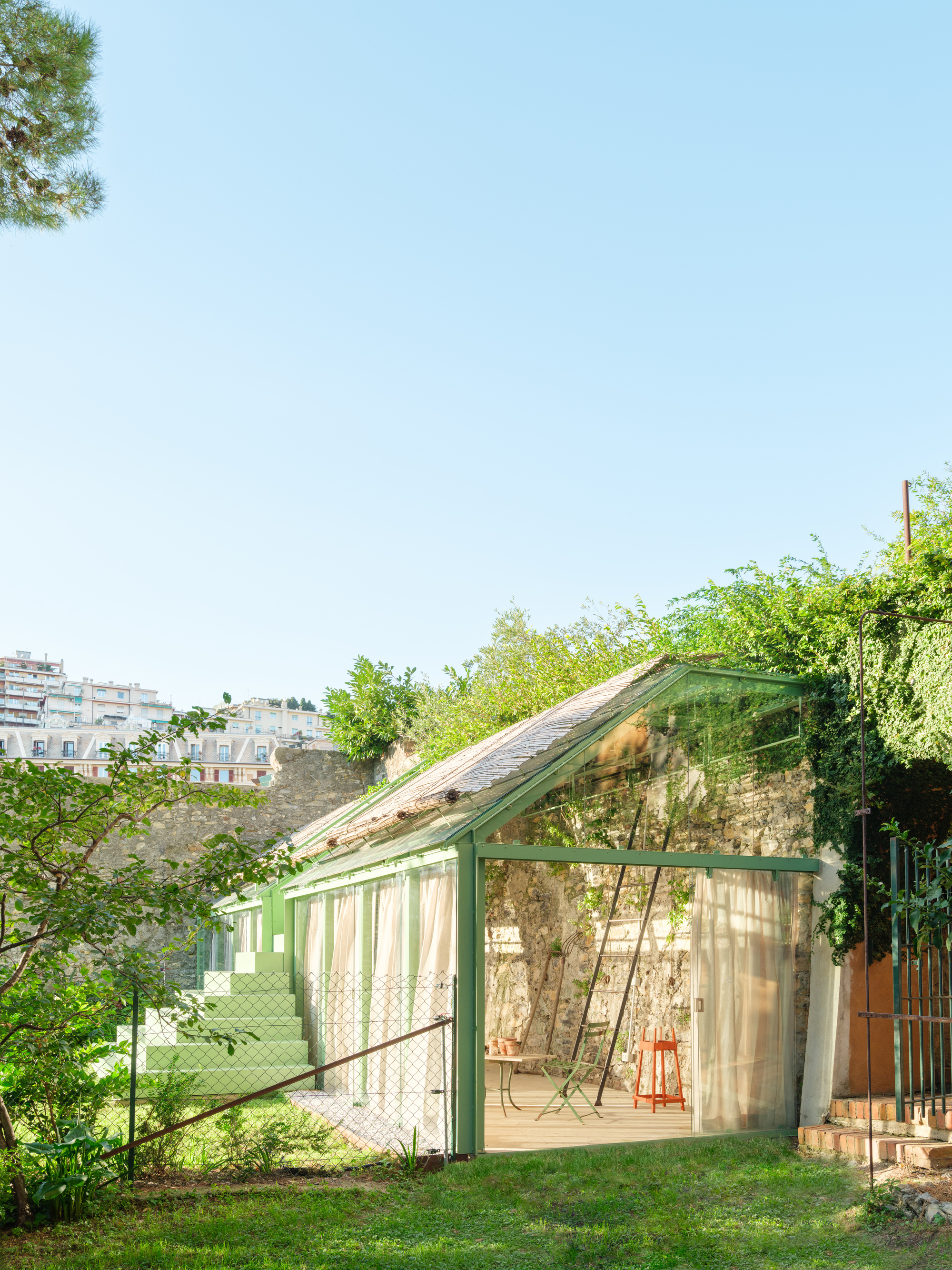 Exterior of the green-framed glass shed against the old stone wall, with curtains, stepped platform and lawn under a clear blue sky