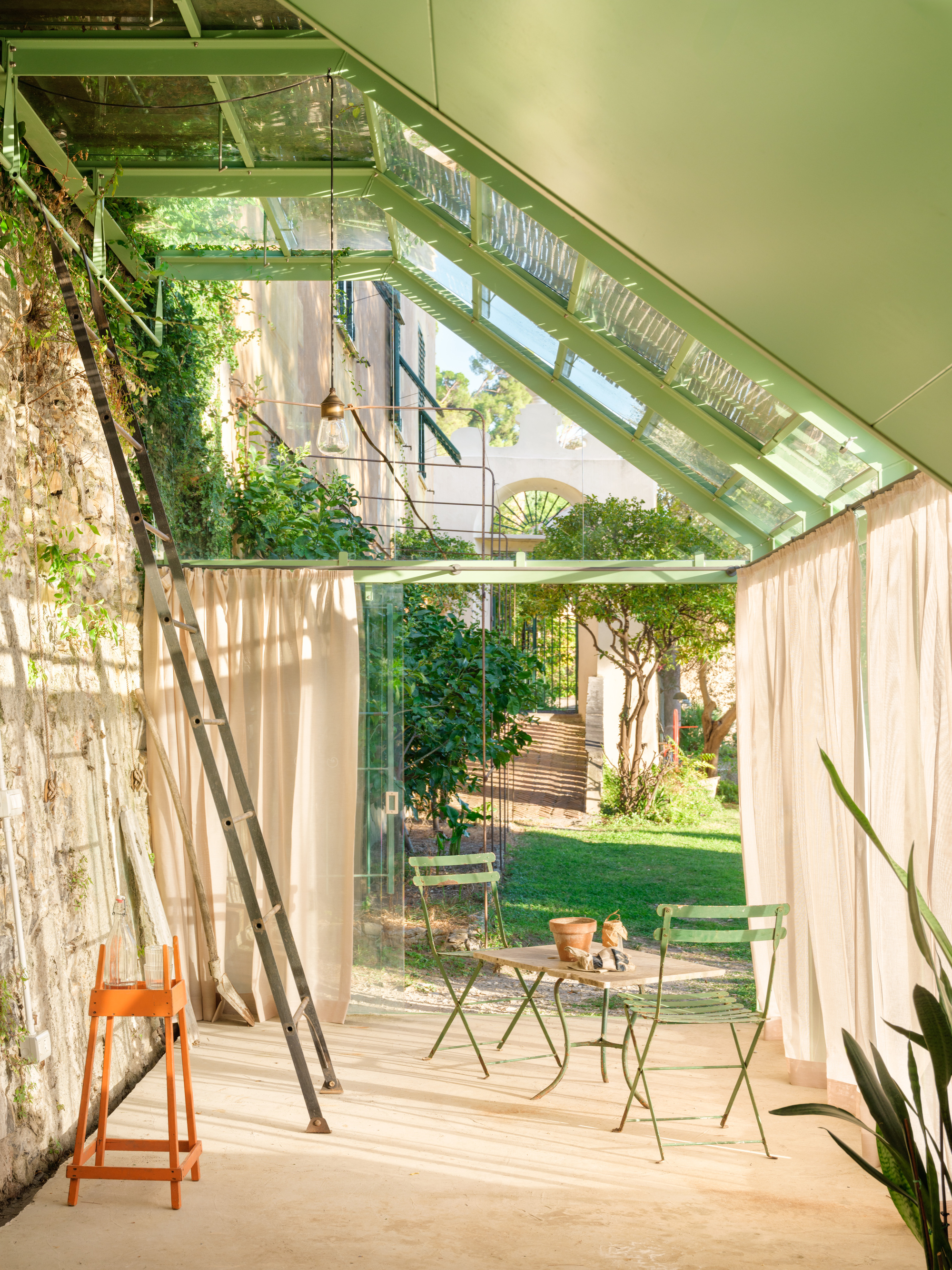 View along the covered interior toward the garden, with a leaning ladder, orange stool and bistro table in late afternoon light