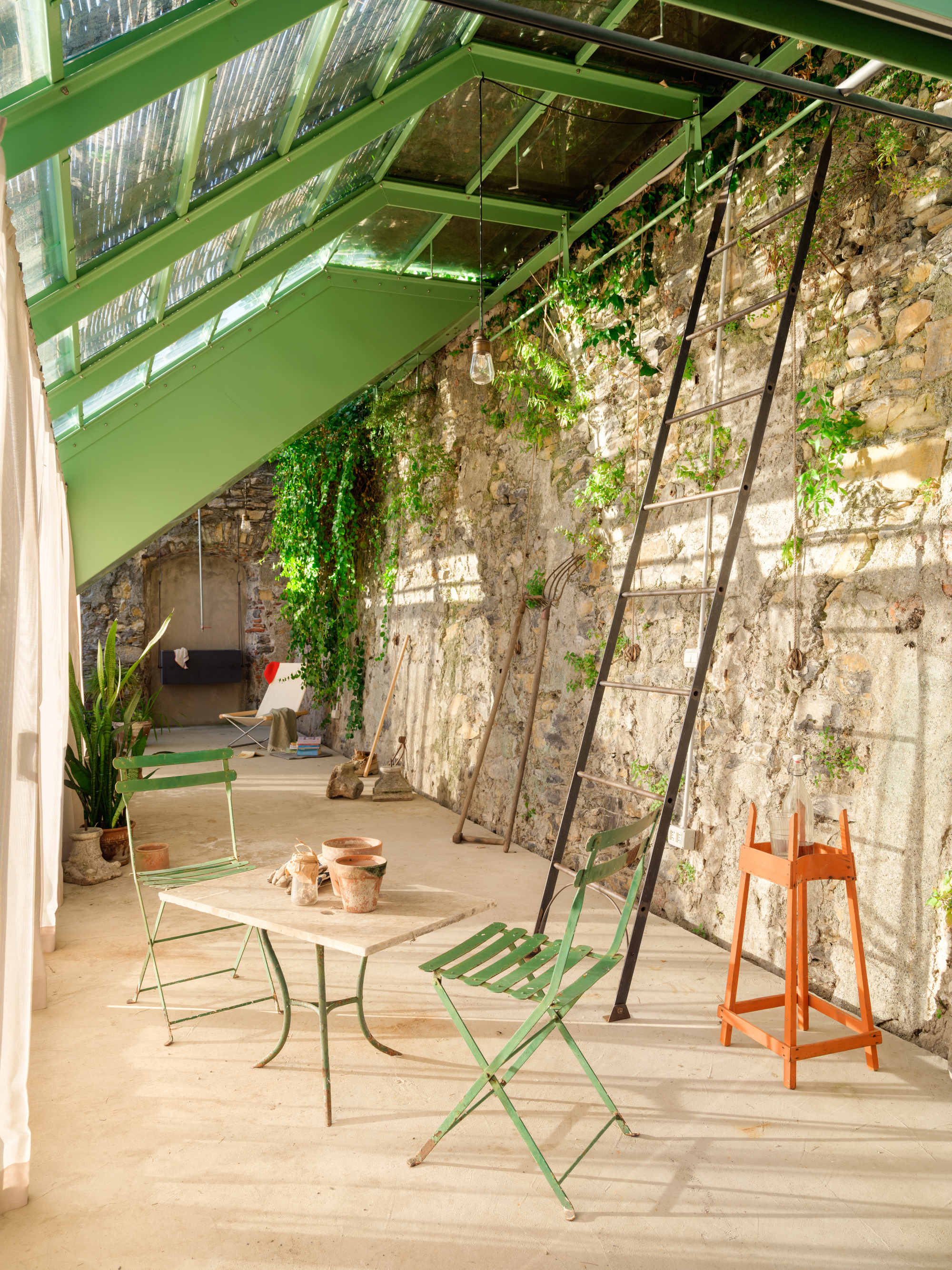 Greenhouse interior with green steel glazing frames, folding bistro chairs and a black ladder against an ivy-draped stone wall in afternoon sun