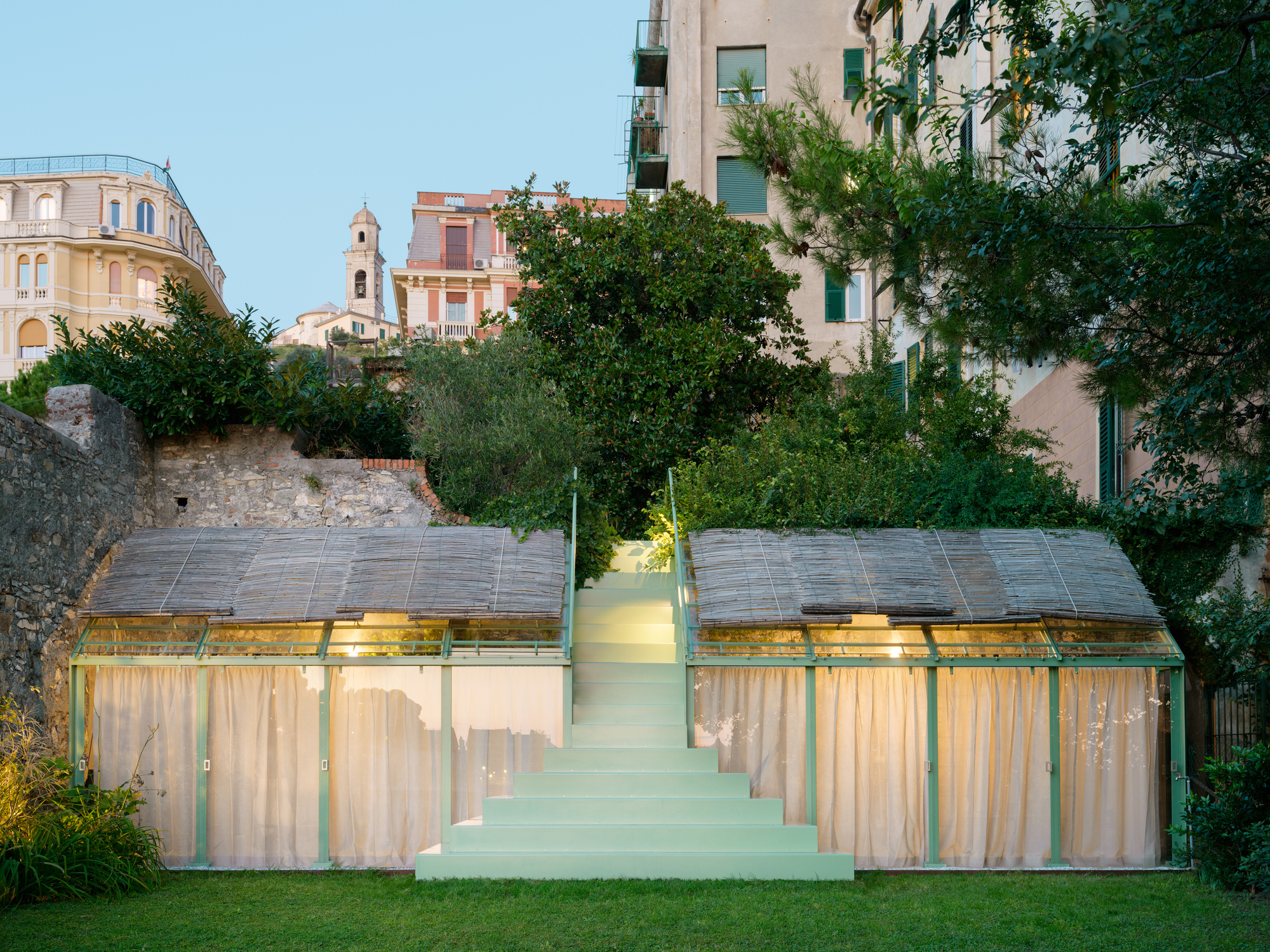 Pale green stepped stair rising between two curtained glass pavilions with bamboo-screen roofs, seen at dusk beneath a hillside of trees