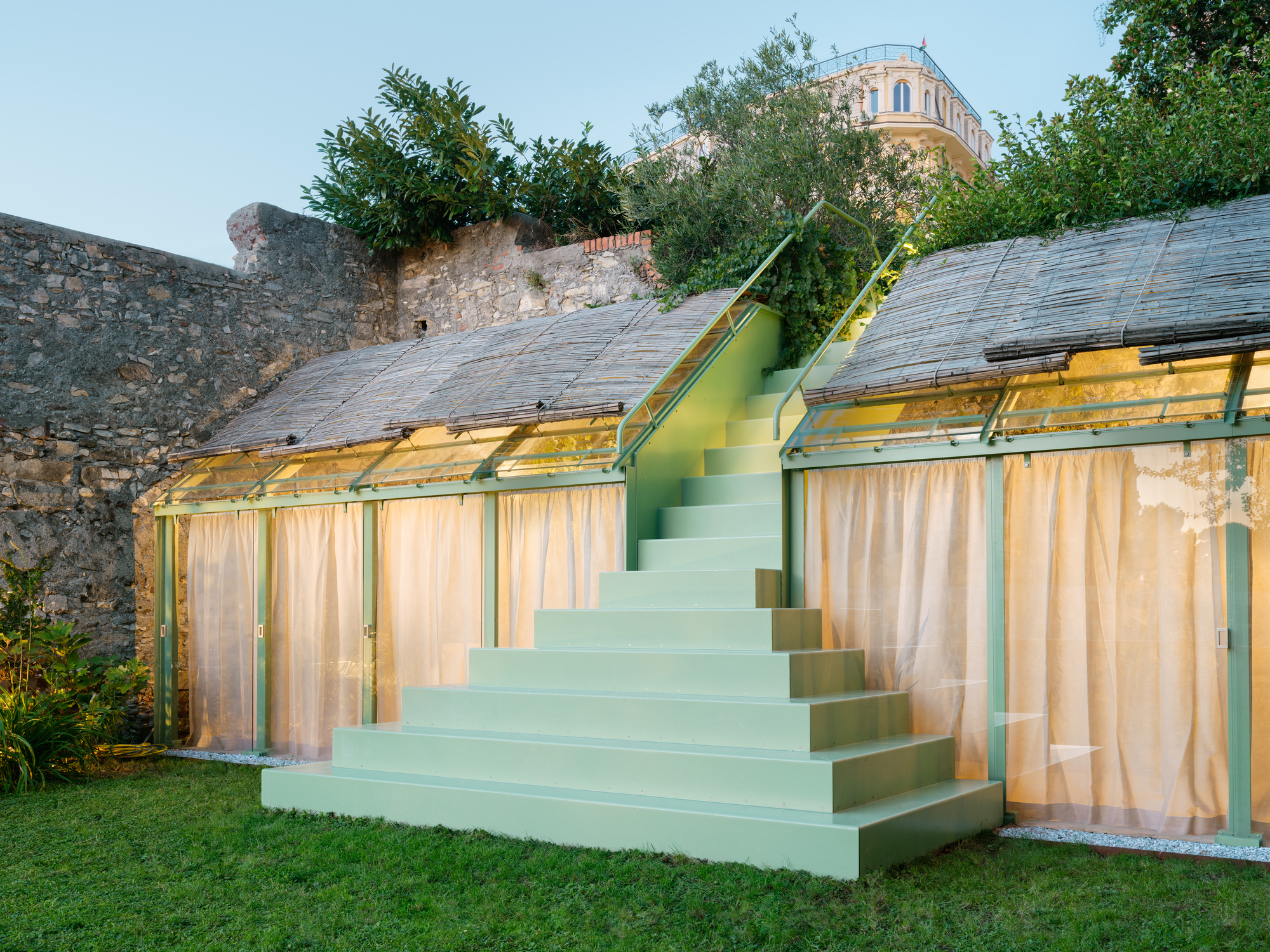 Angled view of the green stair flanked by two glasshouses with reed roofs, set against a stone retaining wall at dusk