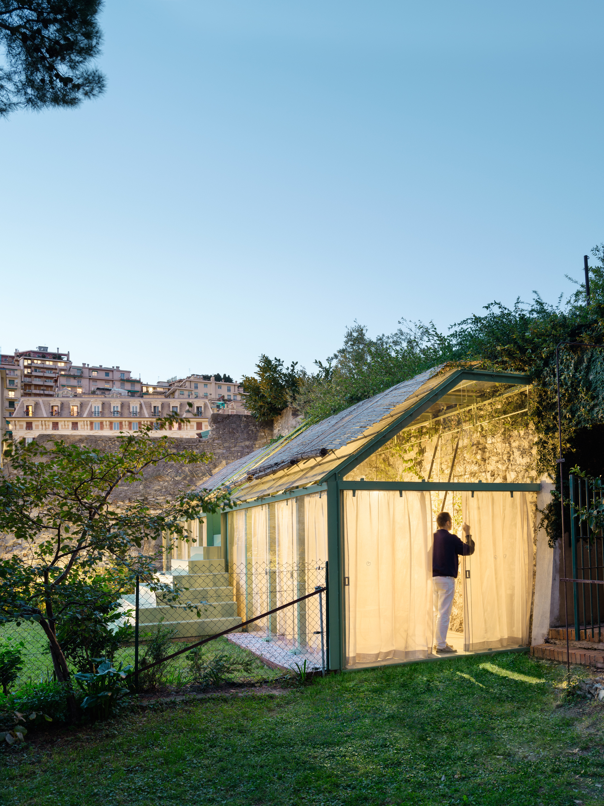 Lit glasshouse with sheer curtains and a man standing inside, set on a lawn beneath hillside apartment buildings at twilight