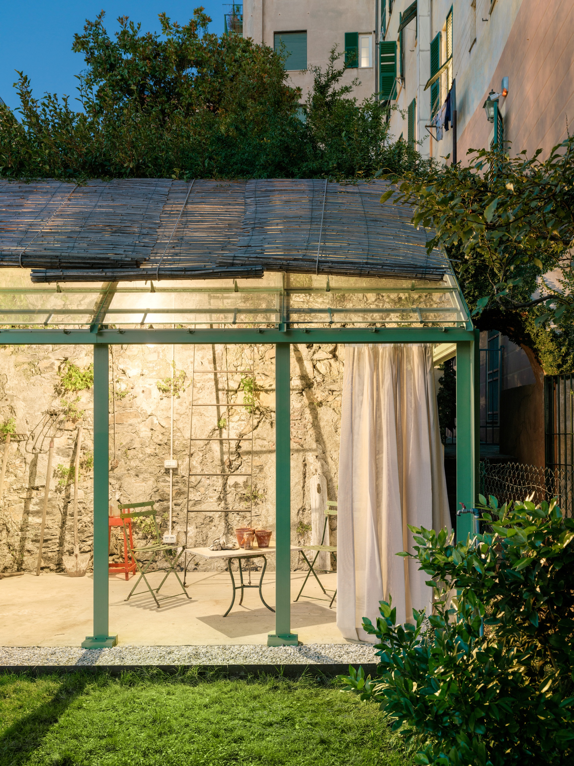 Open side of the glasshouse showing green steel columns, bistro table and chairs, and a ladder against the stone wall at dusk