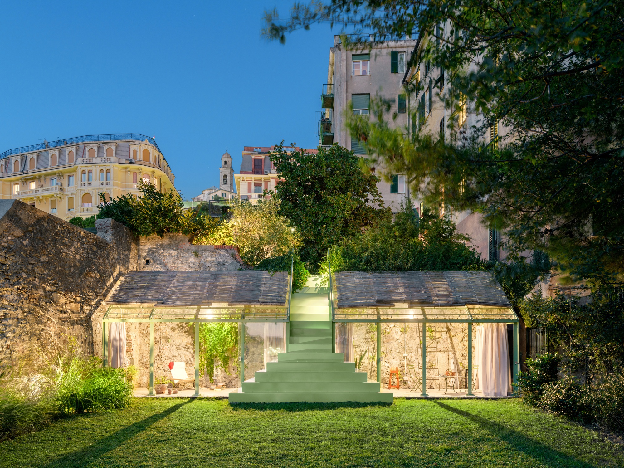 Green stepped stair rising between two glass-walled pavilions with thatched roofs in a lawn garden at dusk, beside a stone retaining wall