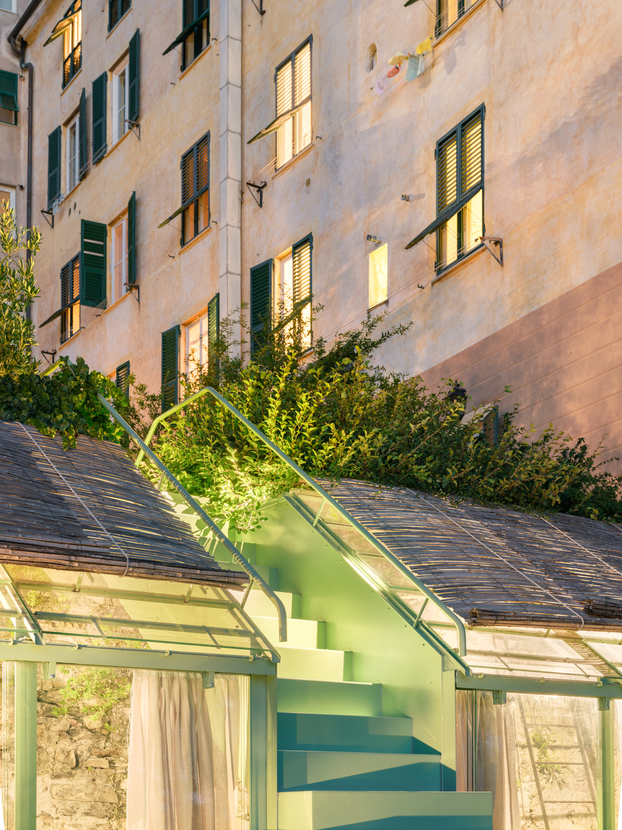 Mint green stair and railing climbing between two reed-covered glazed roofs toward shuttered apartment windows in warm evening light