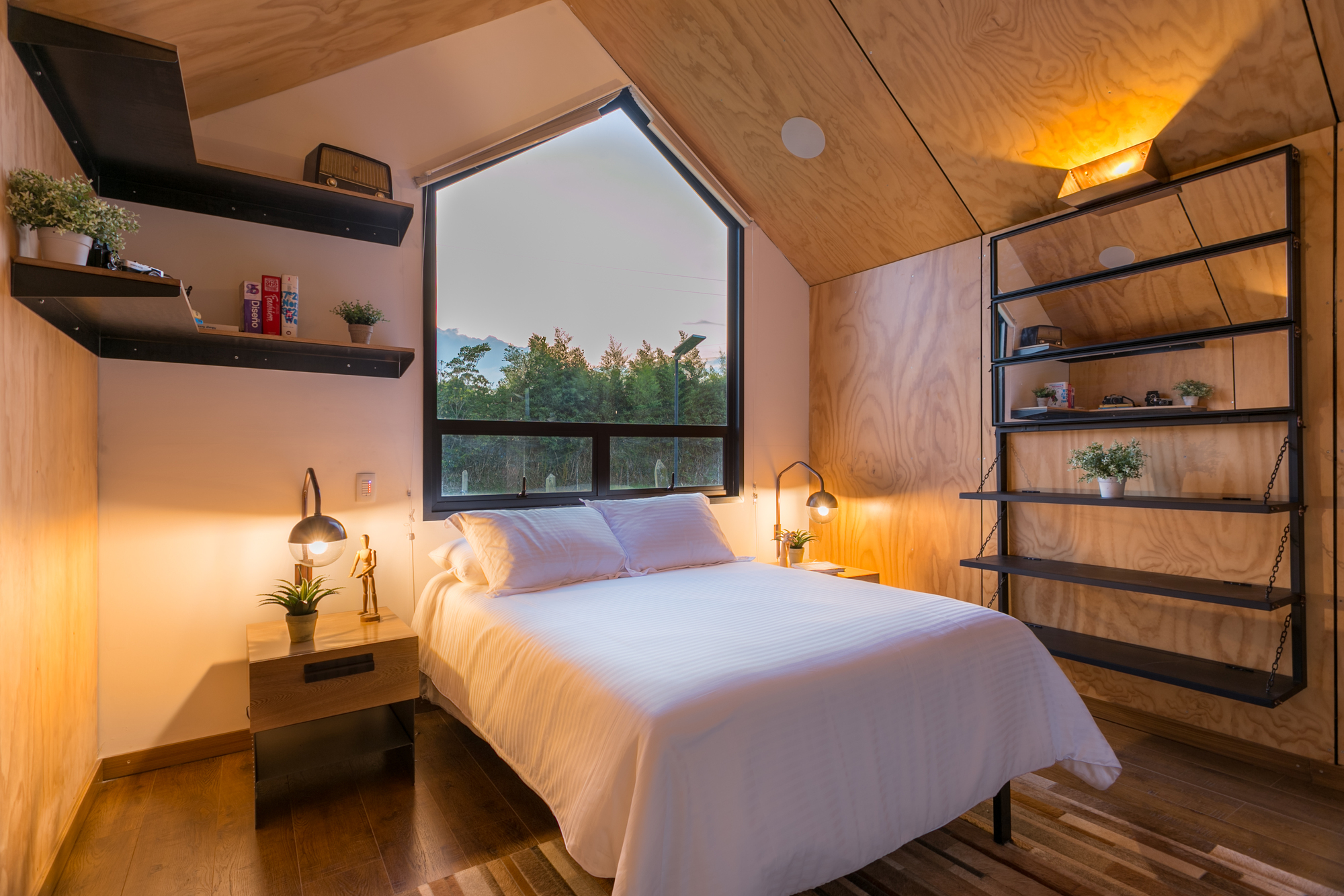Bedroom with a pentagonal window above a white-linen bed, plywood walls and ceiling, and black steel shelving lit by warm lamps