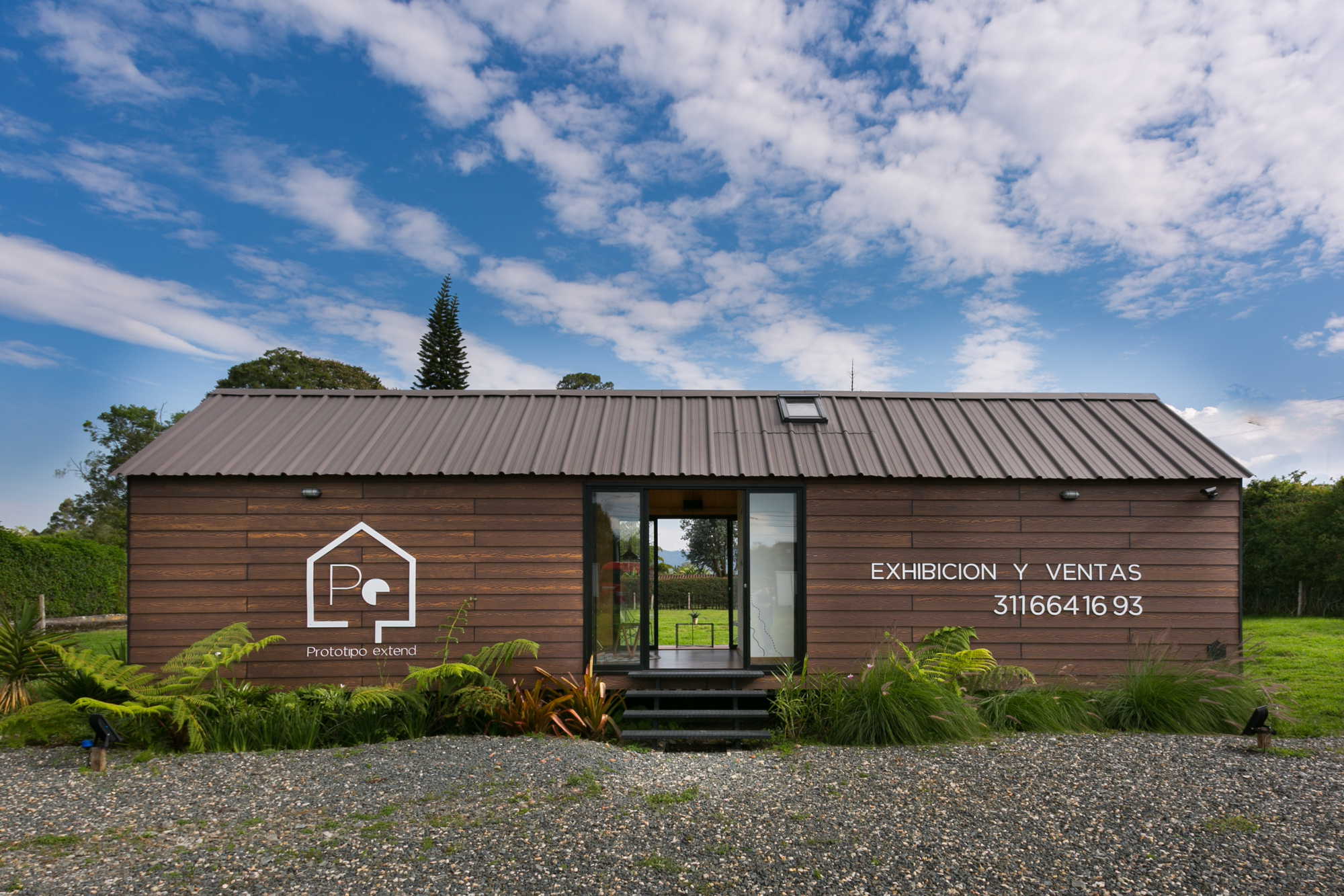 Front elevation of the timber-clad cabin with a standing-seam metal roof and glazed door, set against a blue sky with scattered clouds