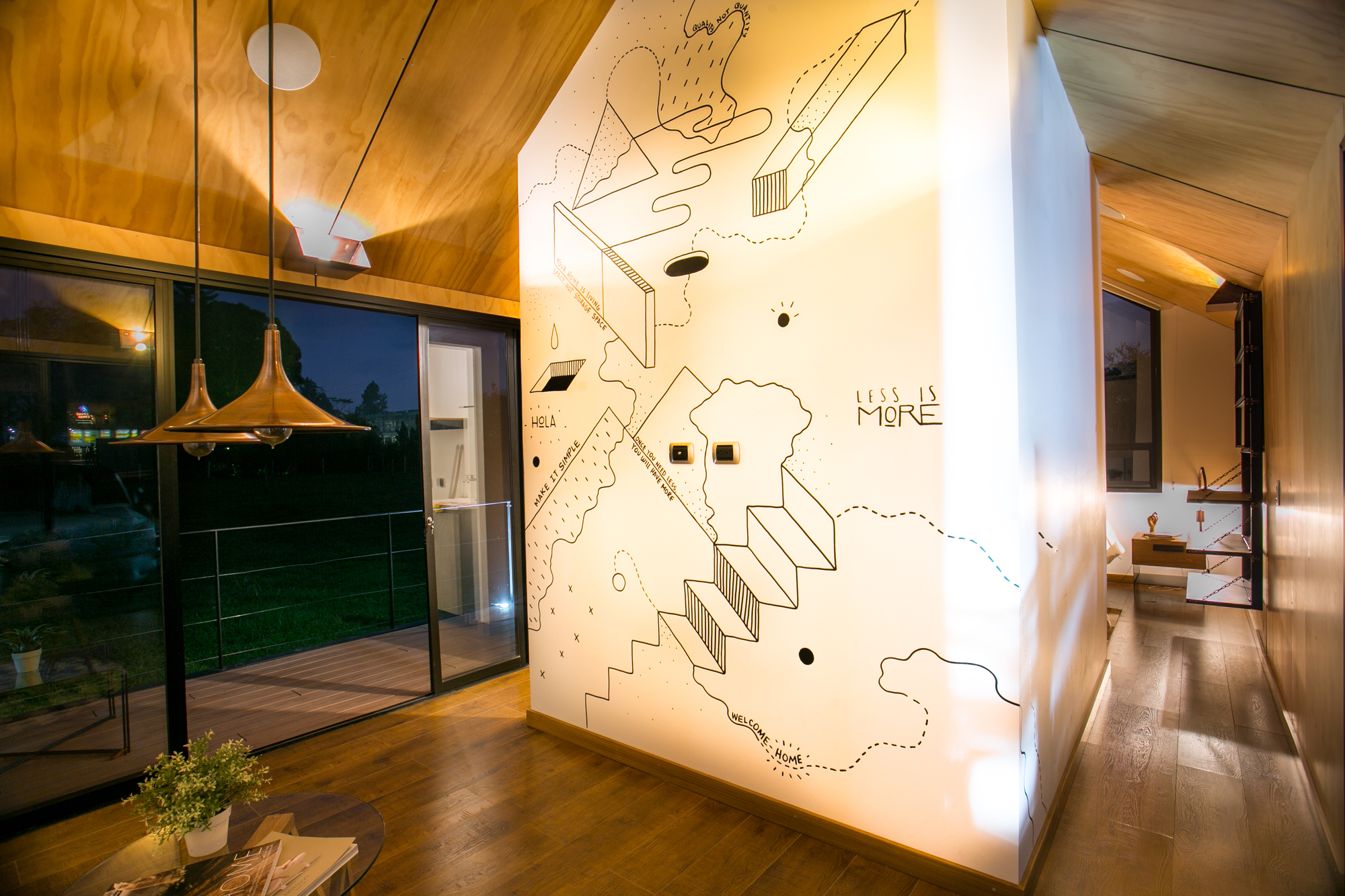 Interior corridor with a white wall covered in black line illustrations, copper pendant lamps and a glazed door to the deck at dusk