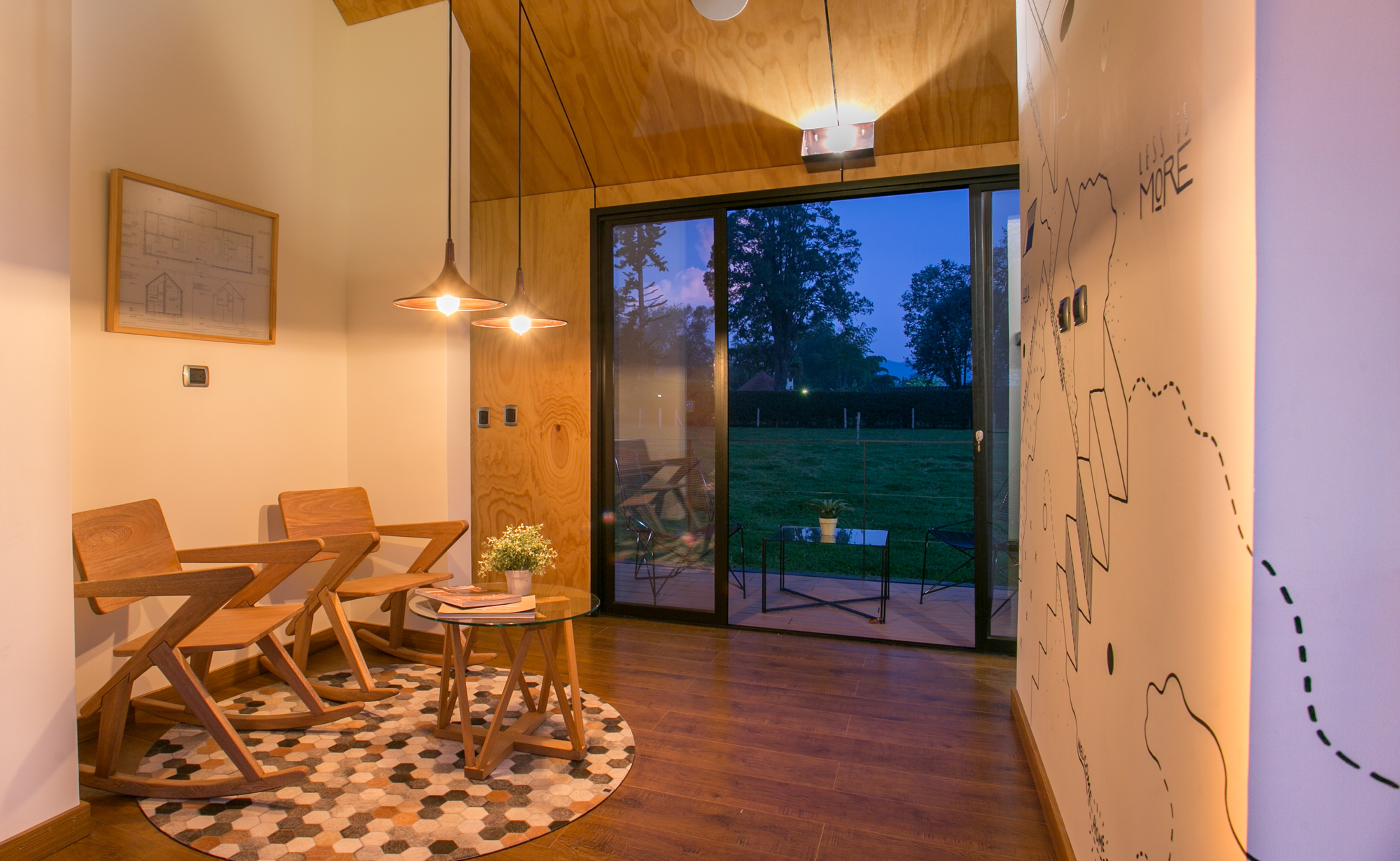 Sitting nook with wooden rocking chairs and a glass side table on a hexagon-patterned rug, facing sliding glass doors at twilight