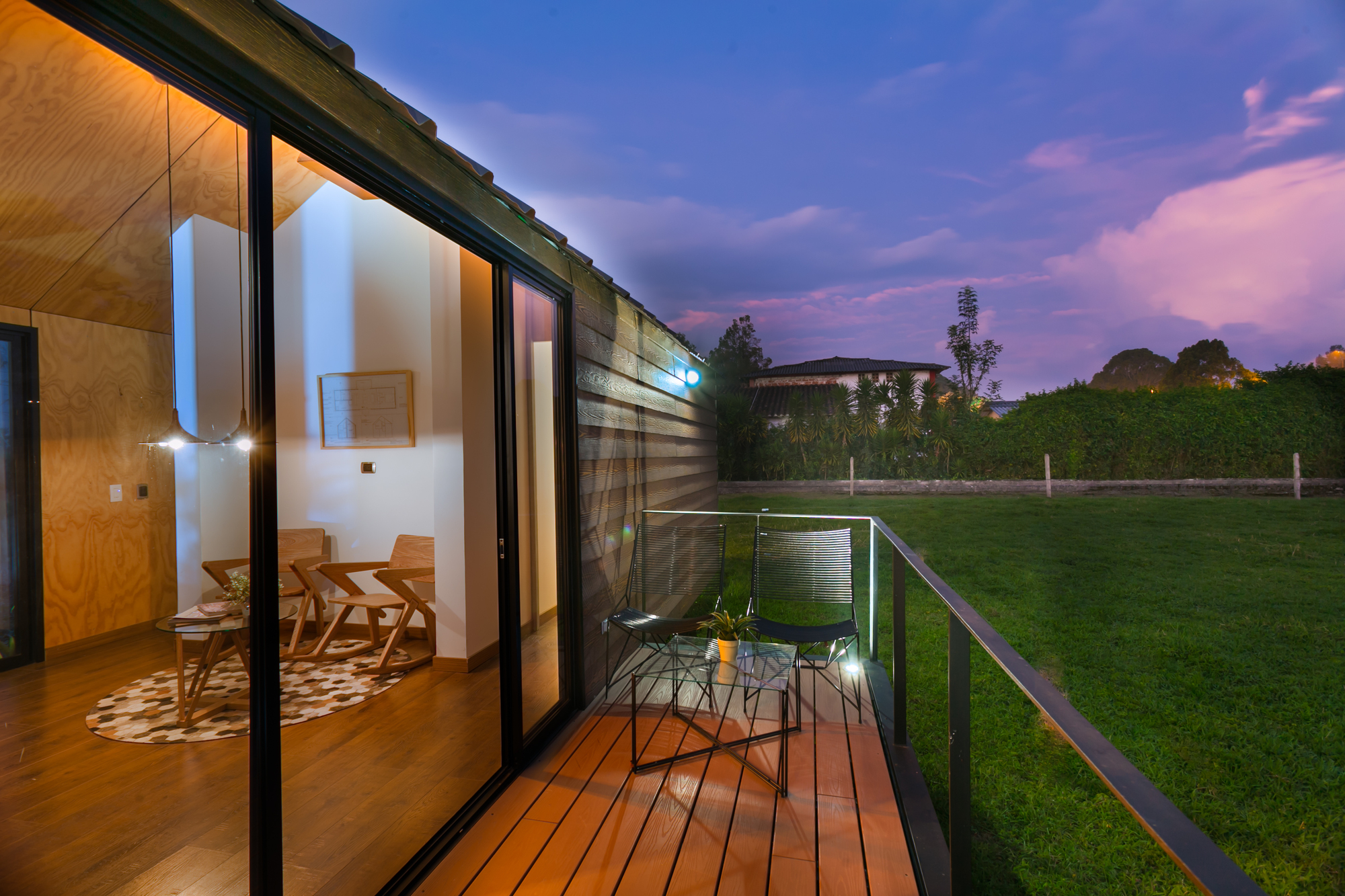 Wooden deck with two metal chairs and a glass table beside the glazed wall, overlooking a lawn under a pink and purple dusk sky