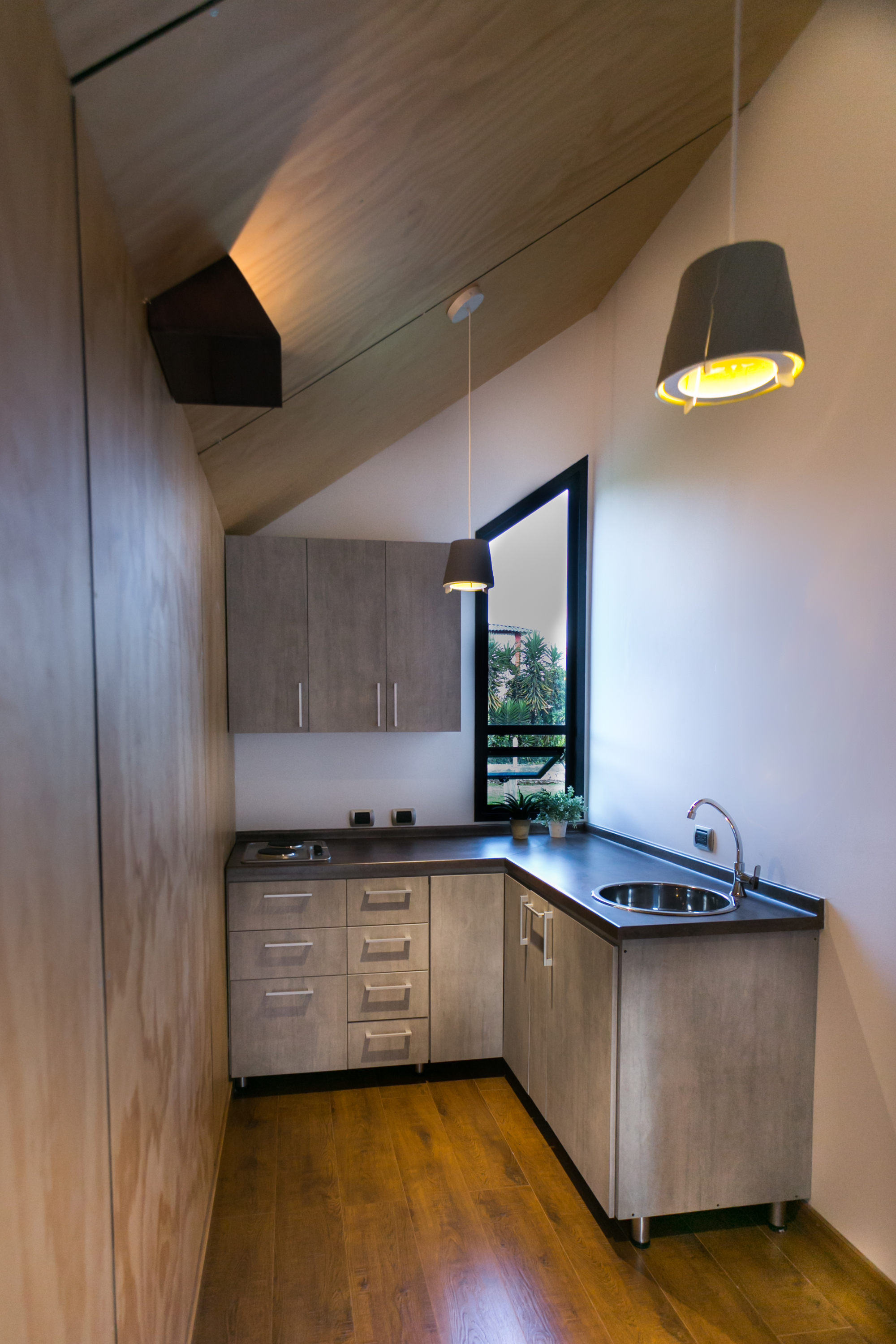 Narrow kitchenette with grey cabinetry, steel sink and a pitched plywood ceiling, lit by a pendant lamp and a small angled window