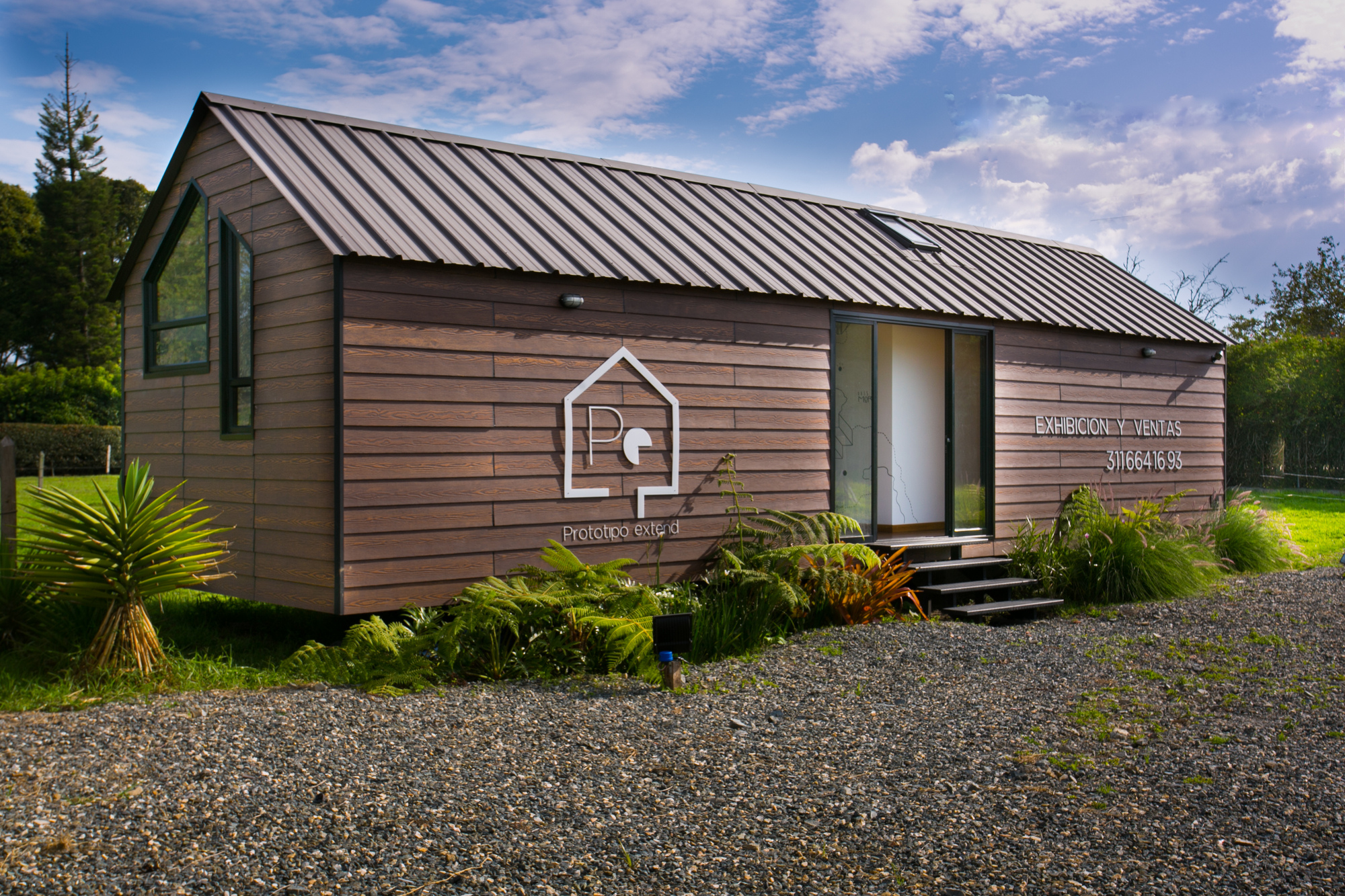Timber-clad cabin with a dark standing-seam metal roof, fern planting and a gravel forecourt under a blue sky with scattered clouds
