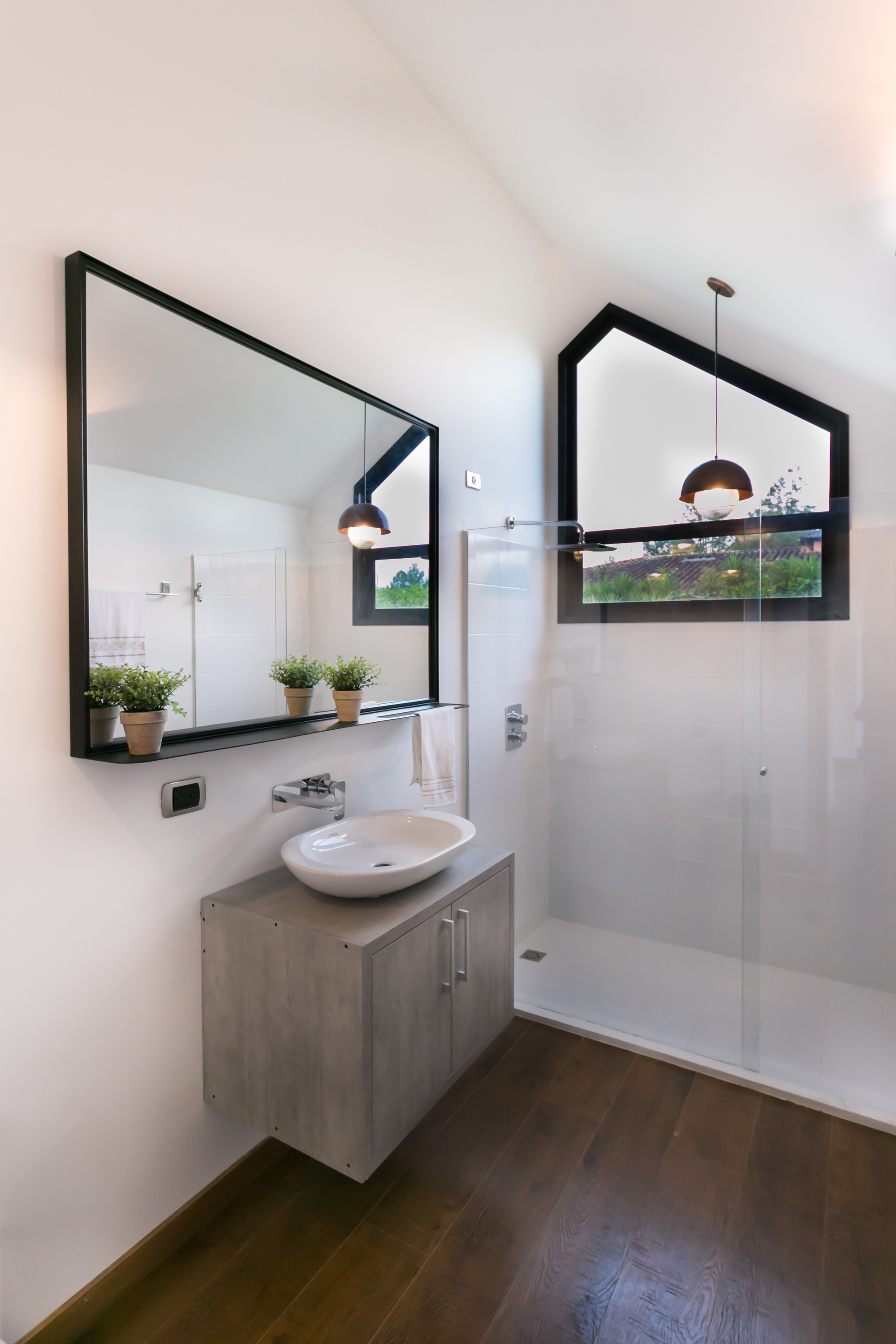 Bathroom with a vessel basin on a floating grey vanity, black-framed mirror and a gable-shaped window above the glass shower