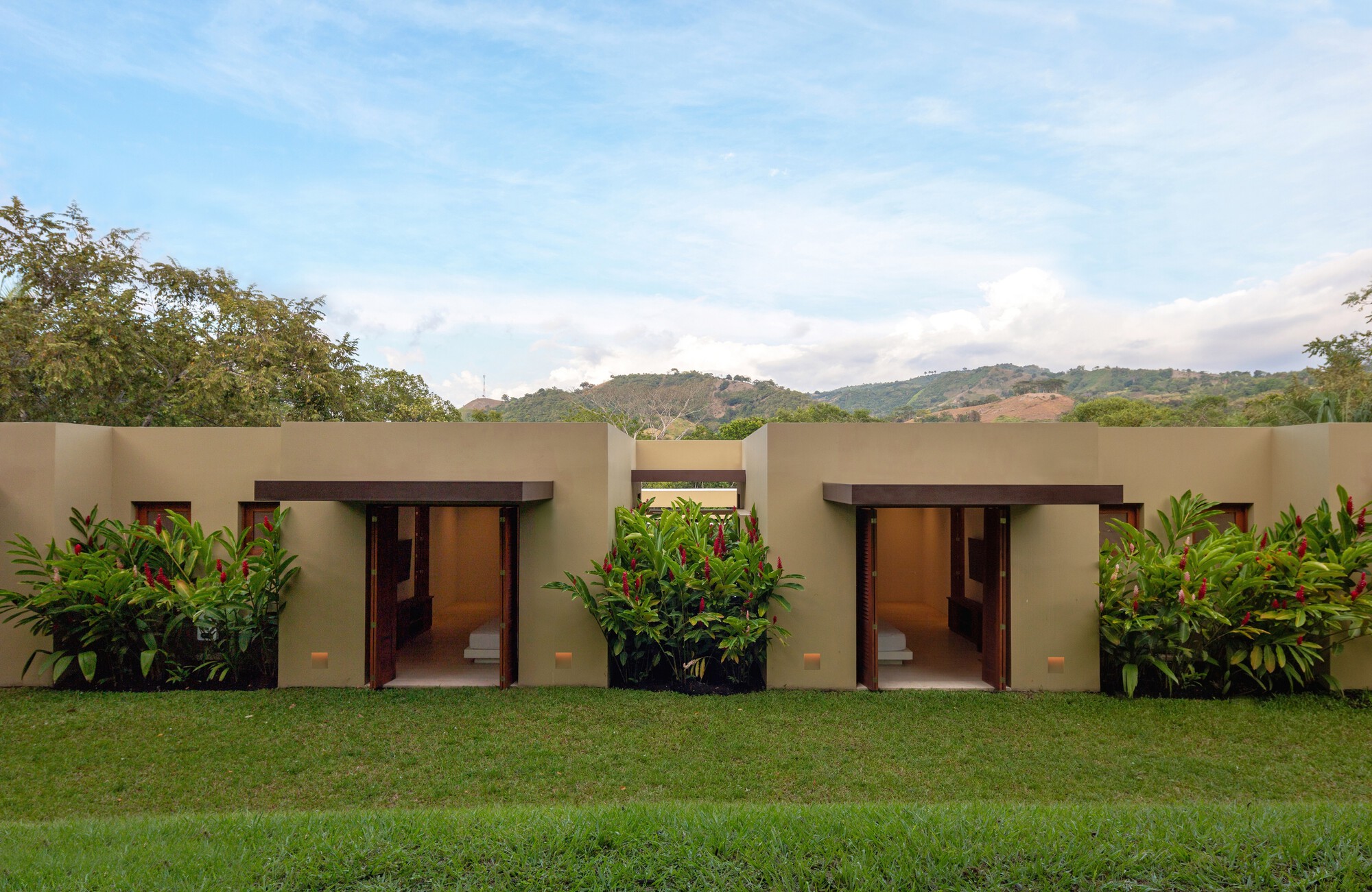 Two open timber doorways in a long ochre wall, flanked by red ginger plants, with hills behind under a partly cloudy sky