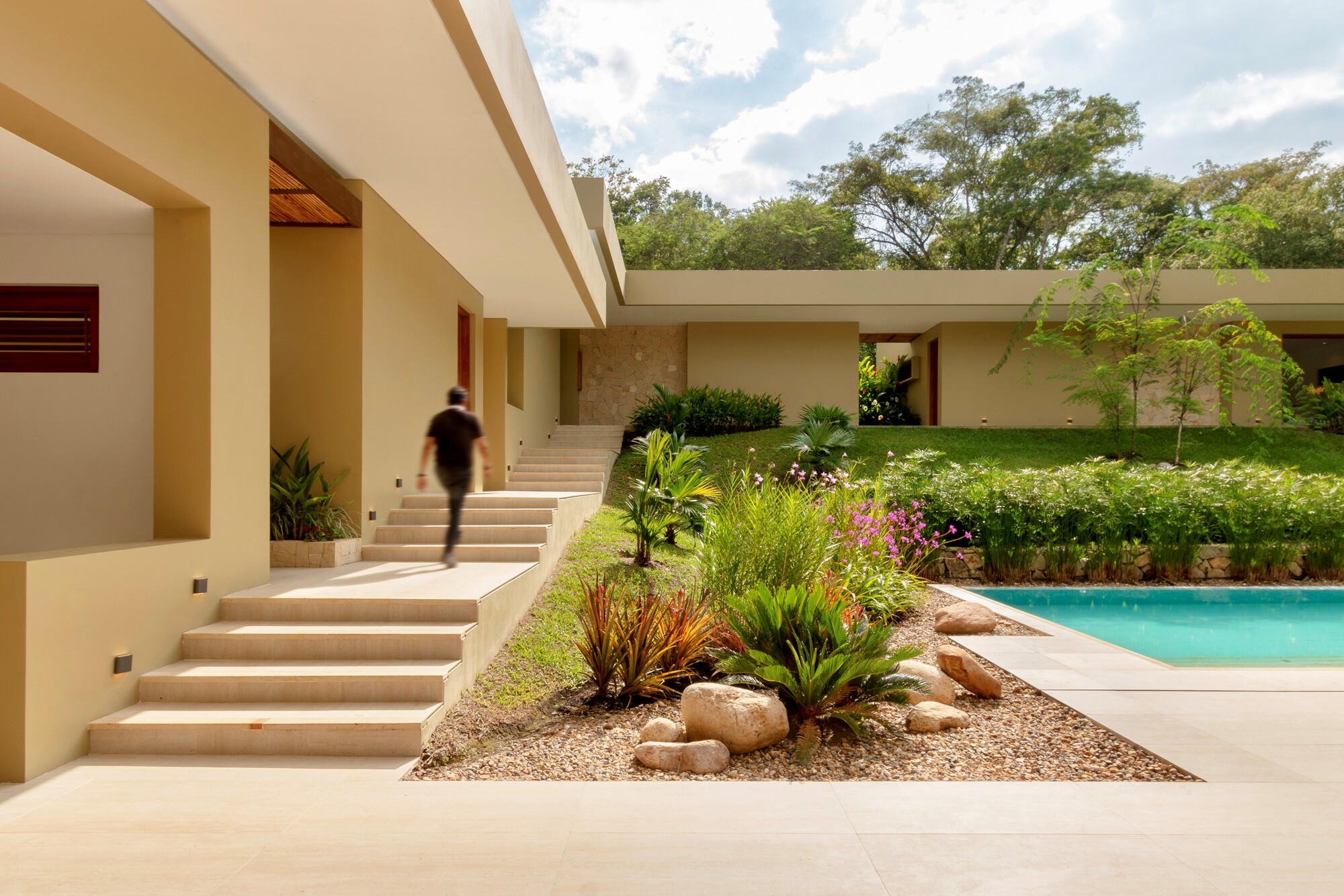 Wide stone steps rising beside a planted gravel bed with boulders and a pool, as a man climbs in daylight