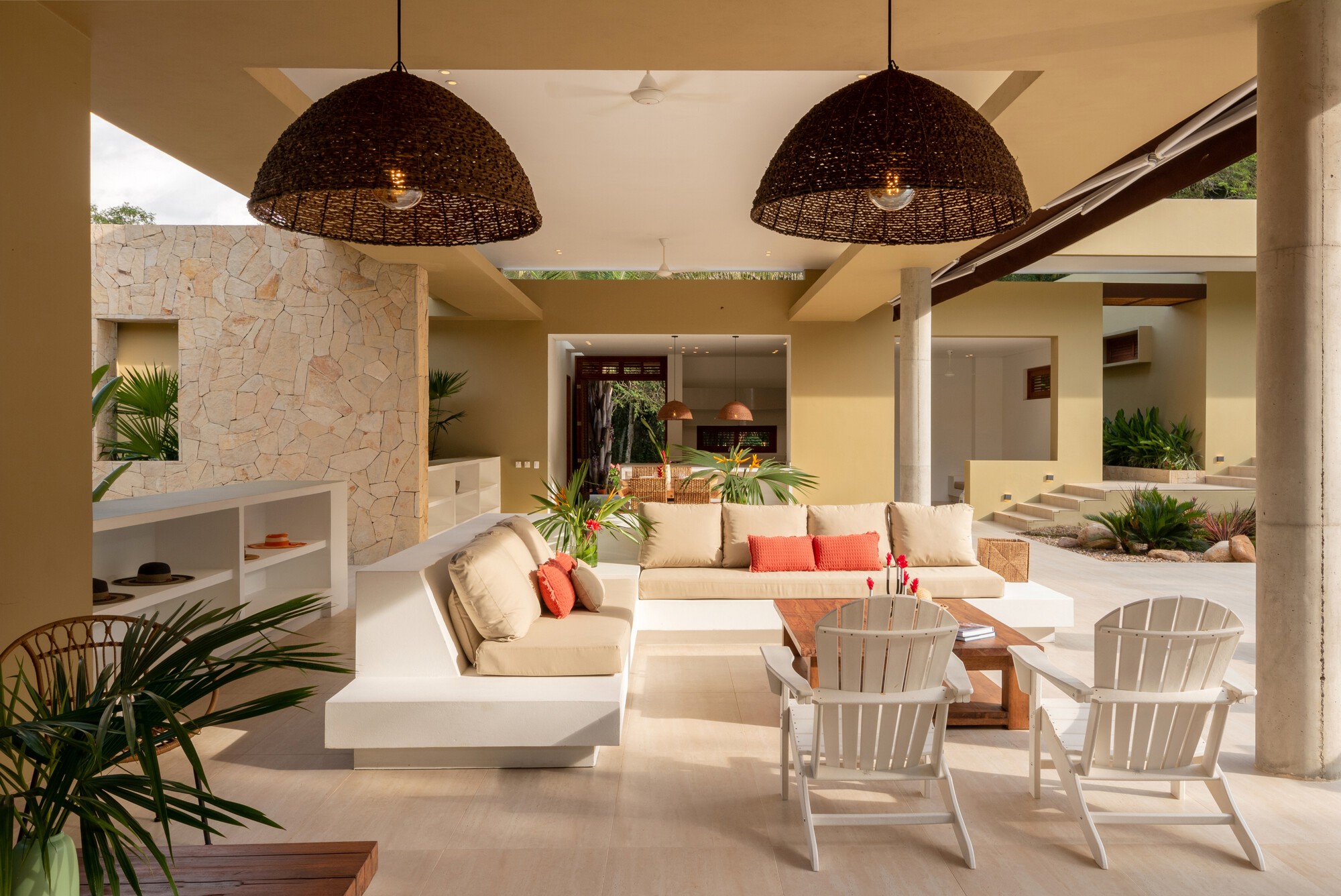 Covered terrace with a white built-in sofa, two woven pendant lamps and slatted white chairs beside a rough stone wall