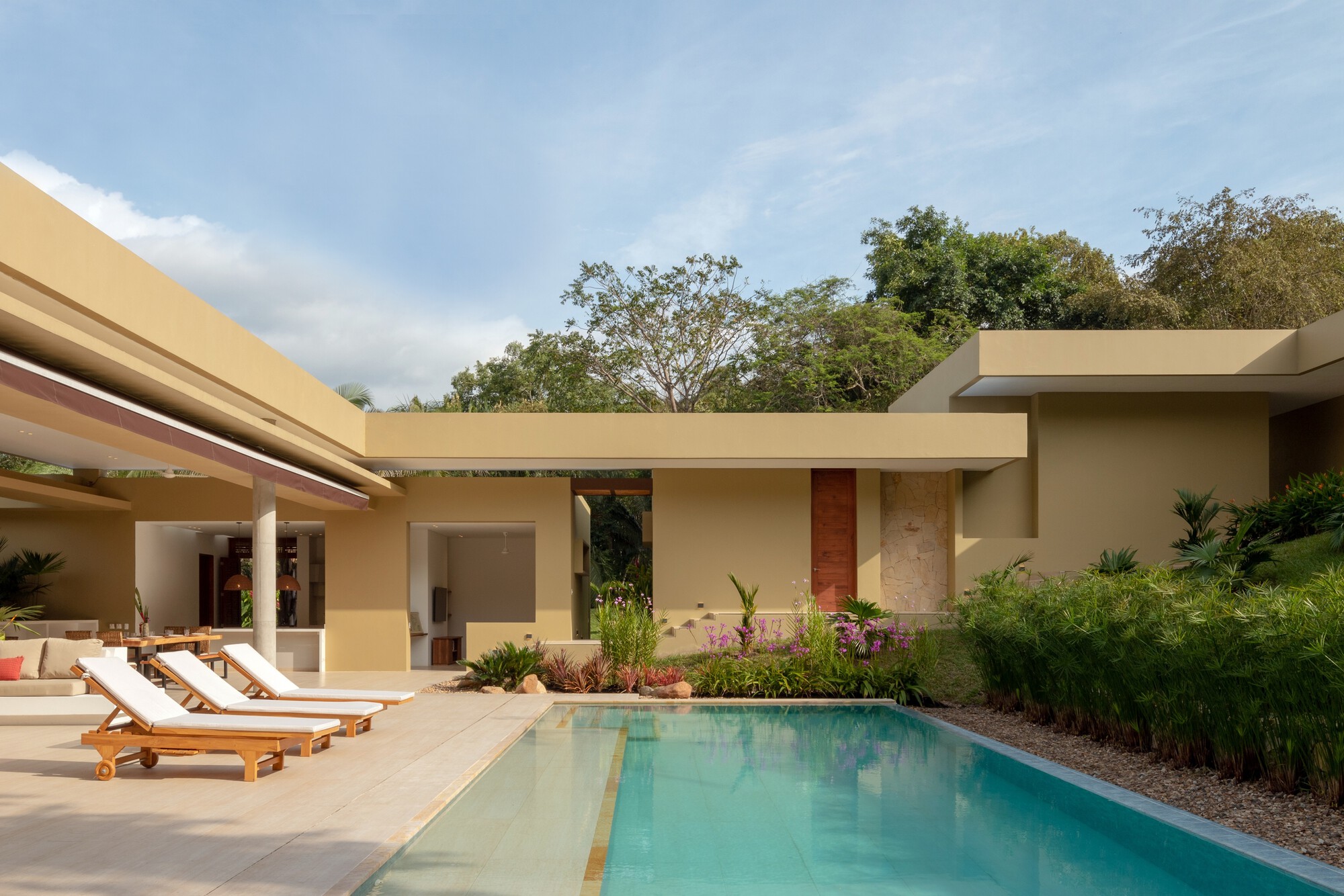 Pool edge with teak loungers in front of flat-roofed ochre volumes, a red timber panel and planting under blue sky