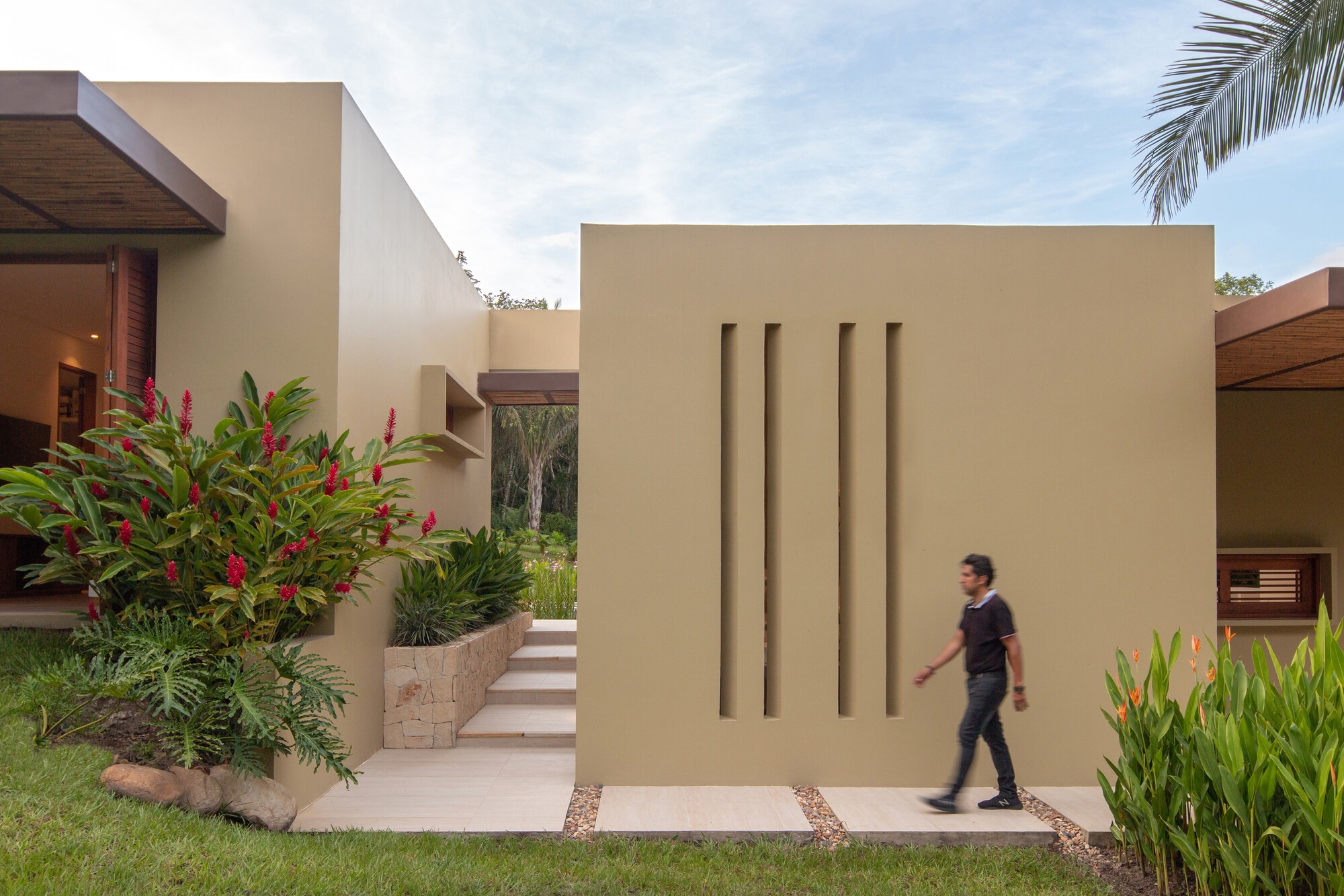 Freestanding ochre wall with four vertical slots as a man walks past, with red ginger and a stone-clad stair behind