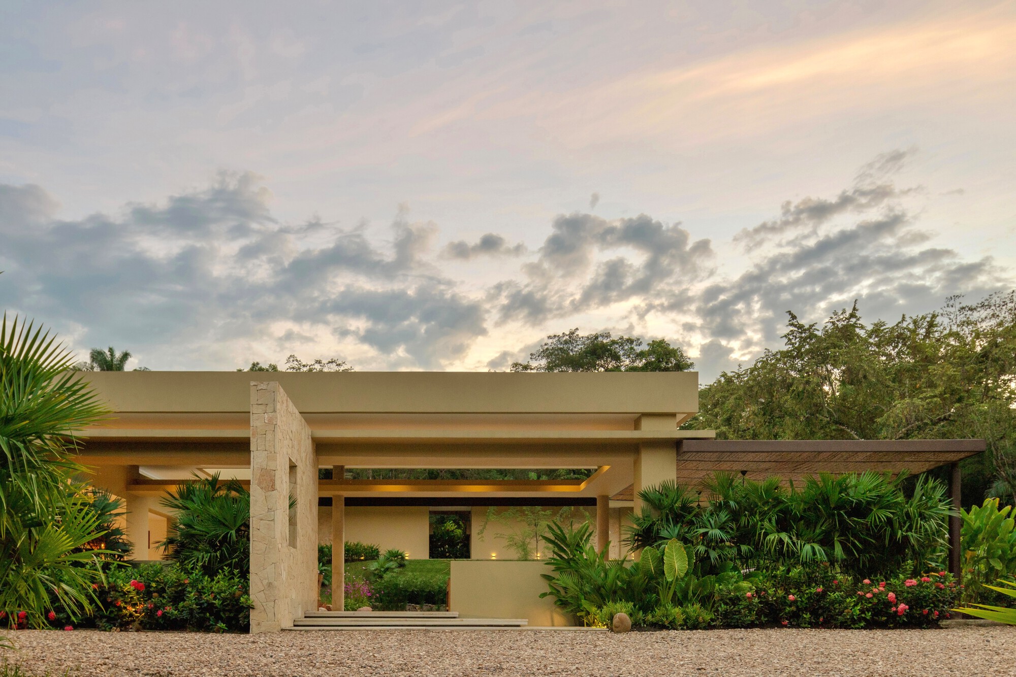 Entrance pavilion with a flat plaster roof and stone pier, framed by palms and red flowering shrubs under a dusk sky