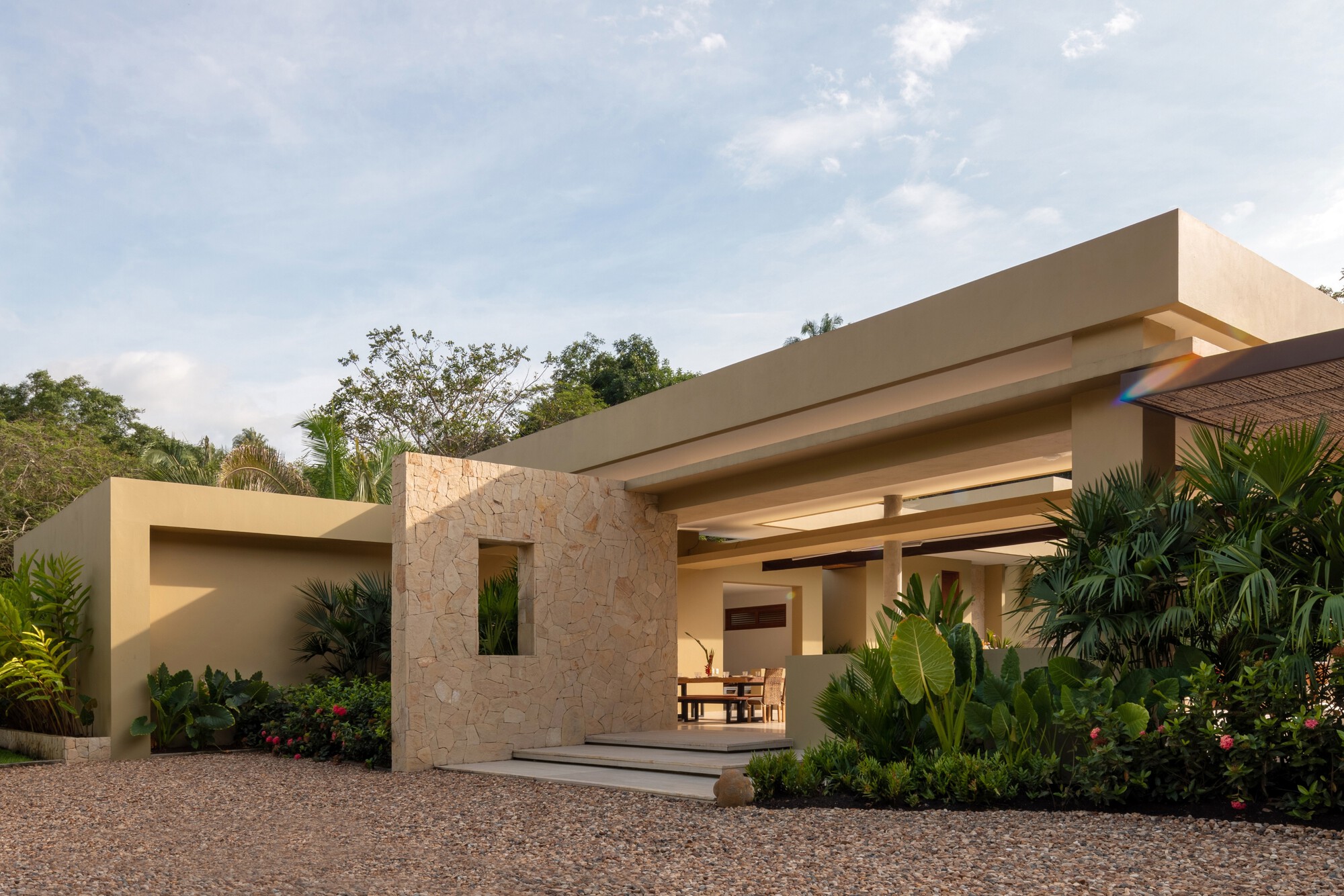 Stone wall with a square window opening beside the entry steps, set in a gravel court with tropical planting in late light