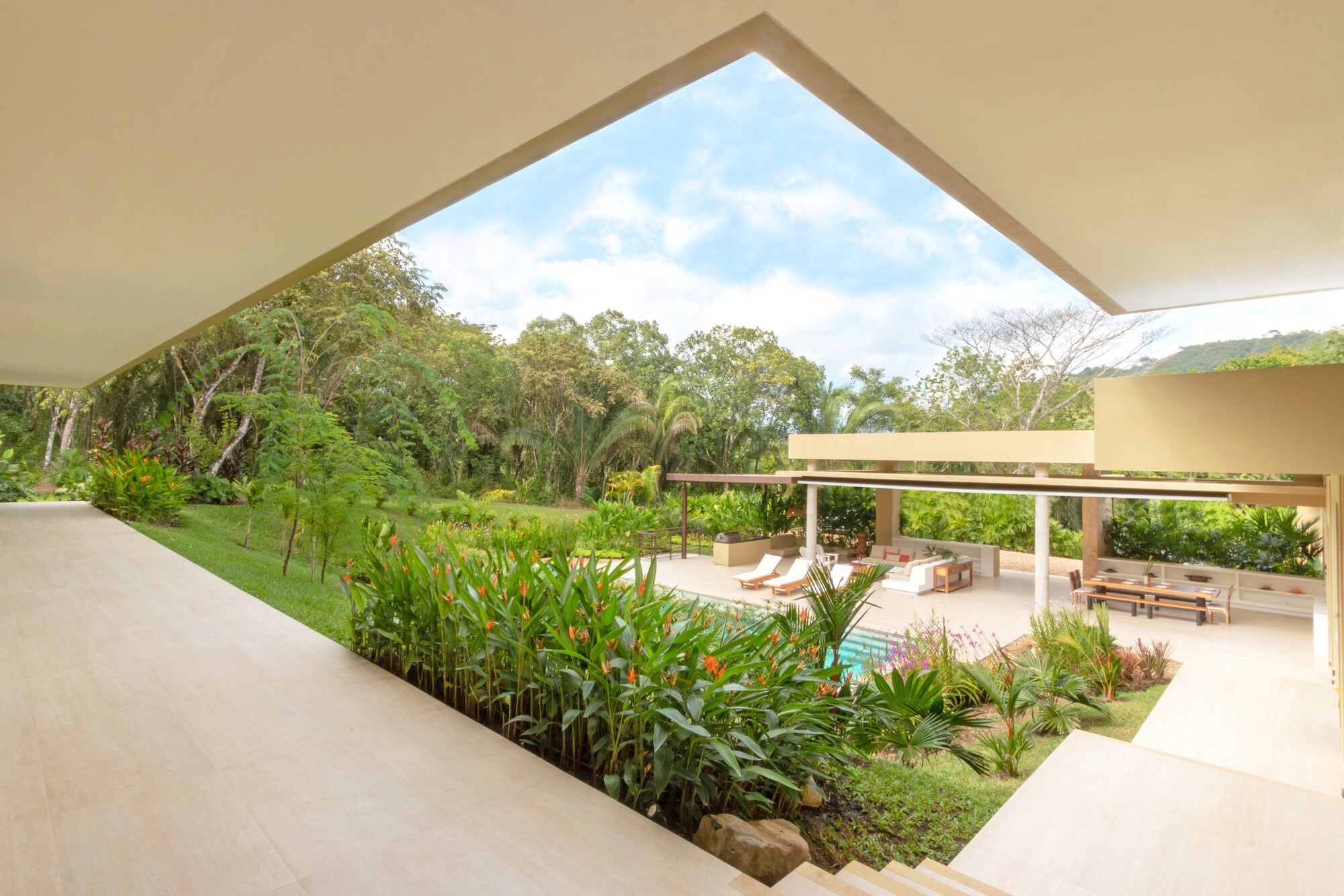View from under a deep roof overhang across a flowering garden to the pool terrace and lounge chairs, with forest beyond