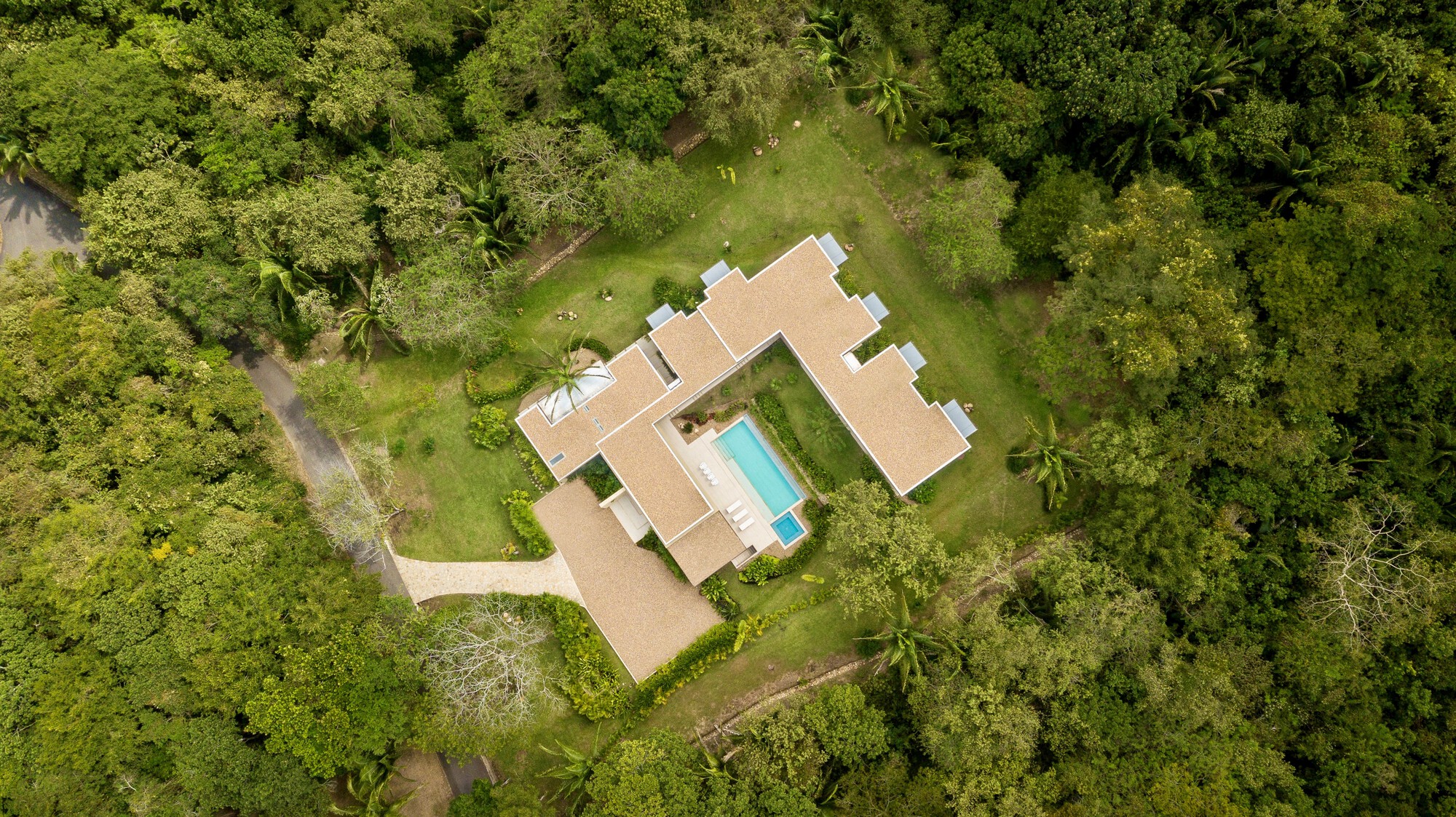 Drone view of the L-shaped house with pale gravel-covered roofs and a turquoise pool, surrounded by dense green forest
