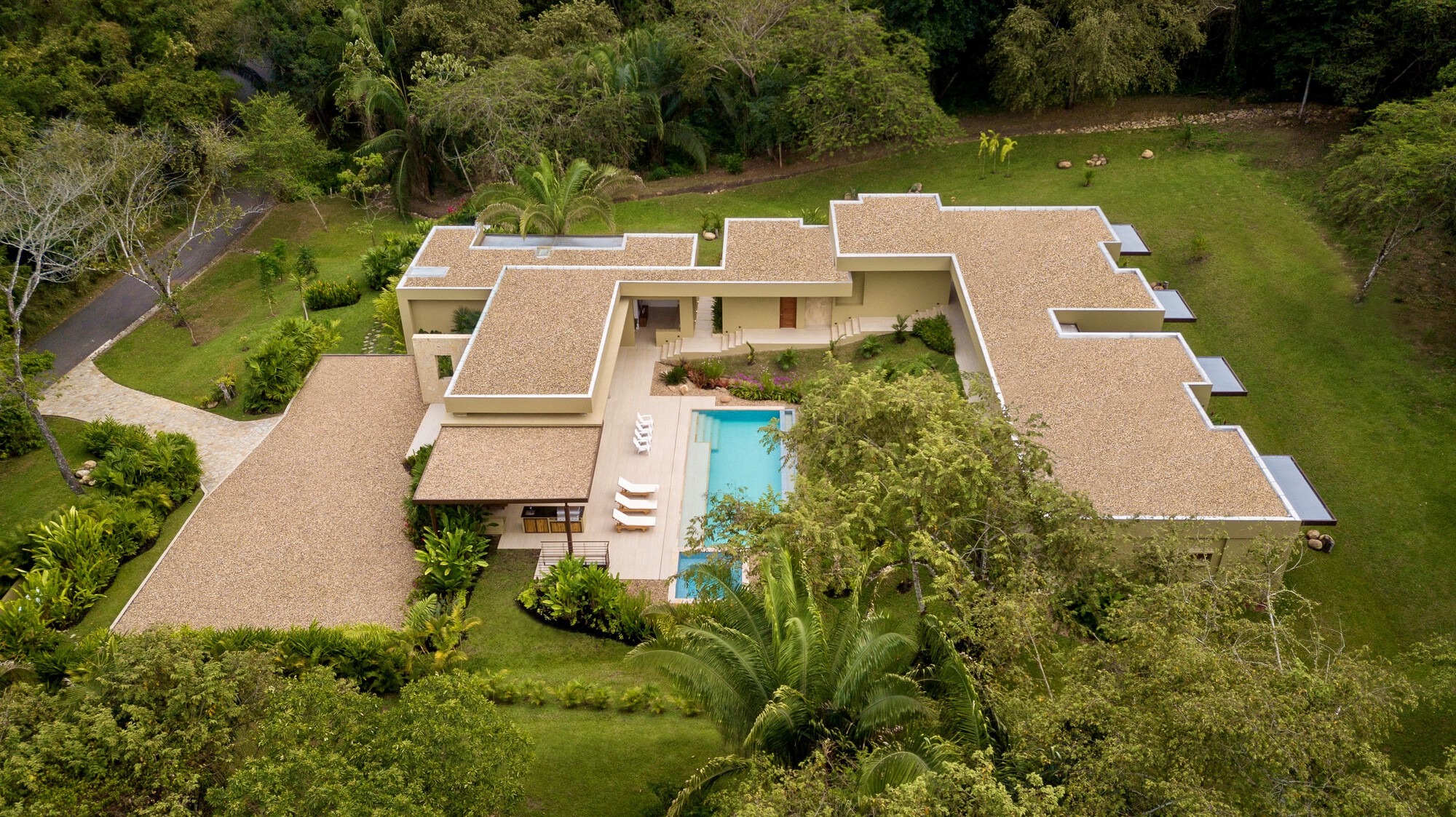 Oblique aerial view of the low gravel-roofed wings wrapping a pool terrace, with a lawn and palms in overcast daylight