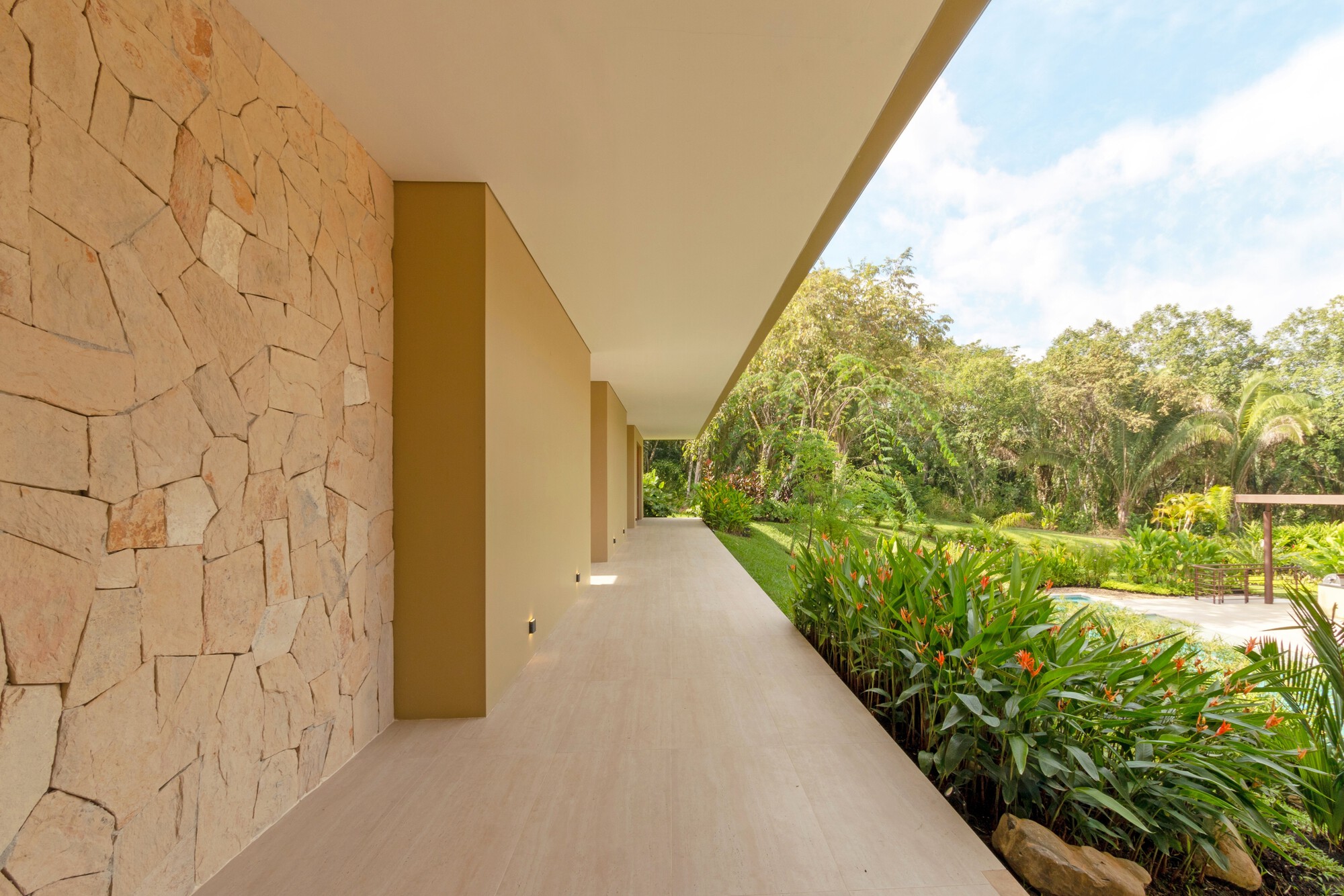 Long covered walkway beside a rough-cut stone wall, with orange heliconia plants and a lawn catching bright afternoon sun