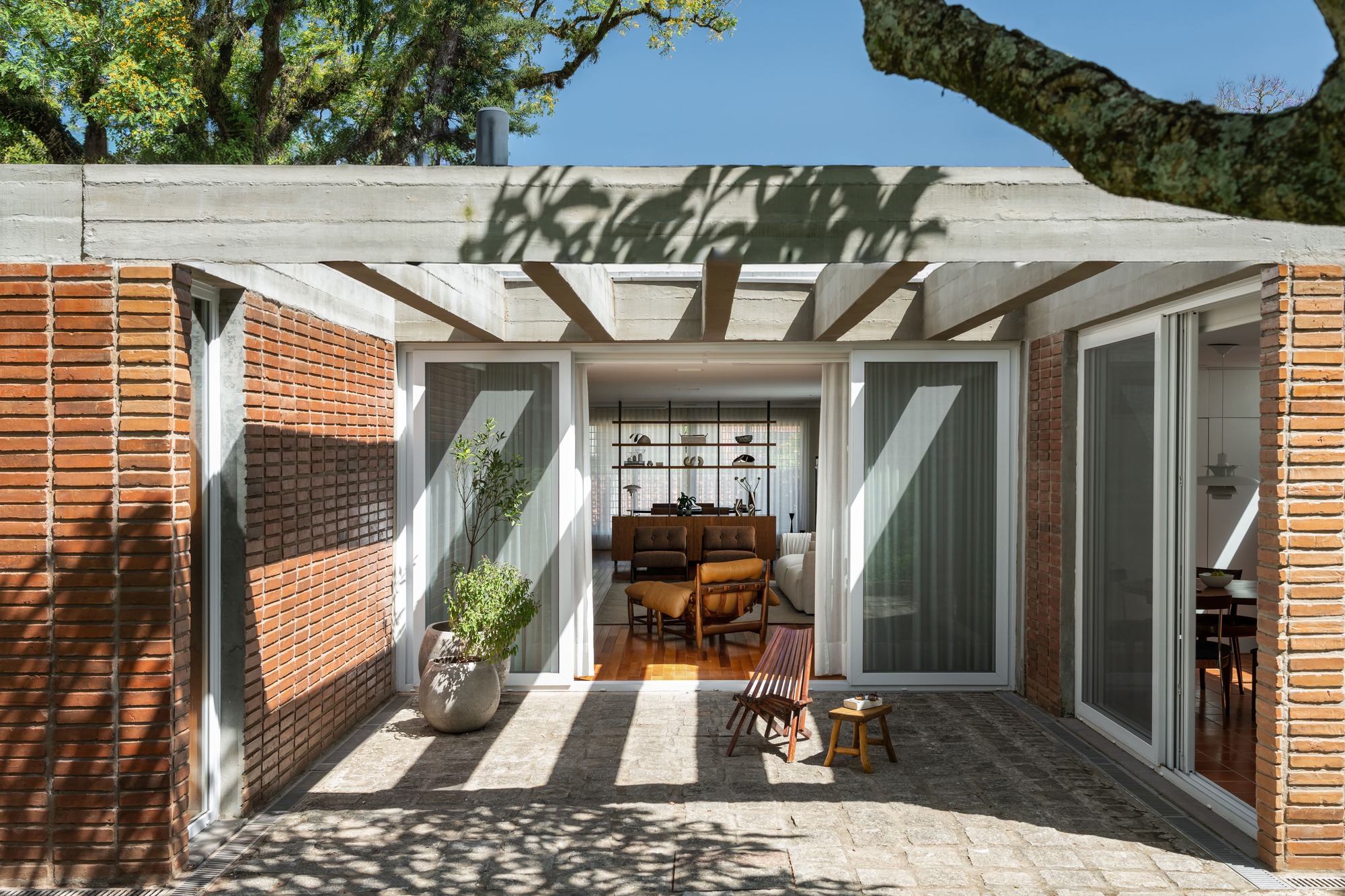 Brick-walled patio with a board-formed concrete beam and pergola, opening through sliding glass doors to a living room, tree shadows on the paving