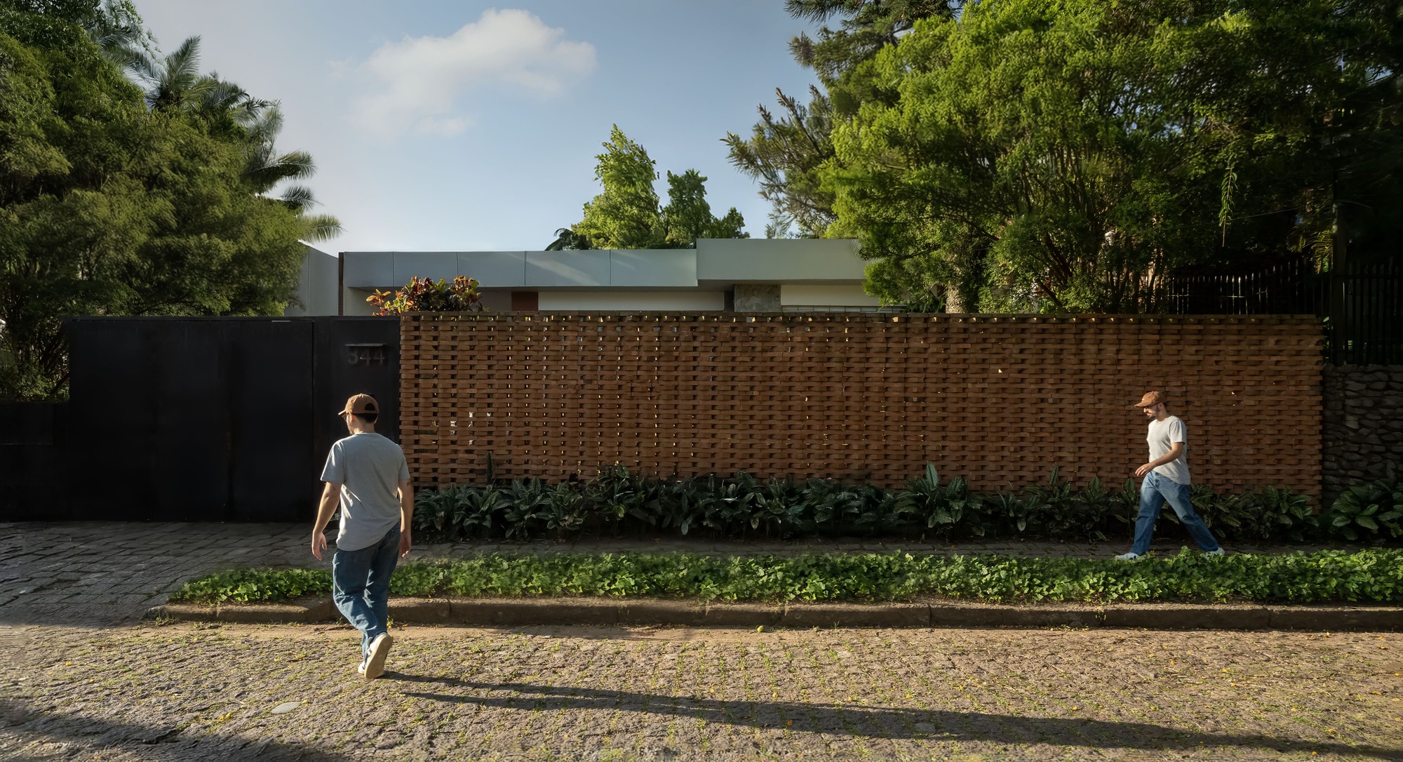 Perforated brick screen wall and black timber gate along a cobbled street, with two men walking past in late afternoon sun