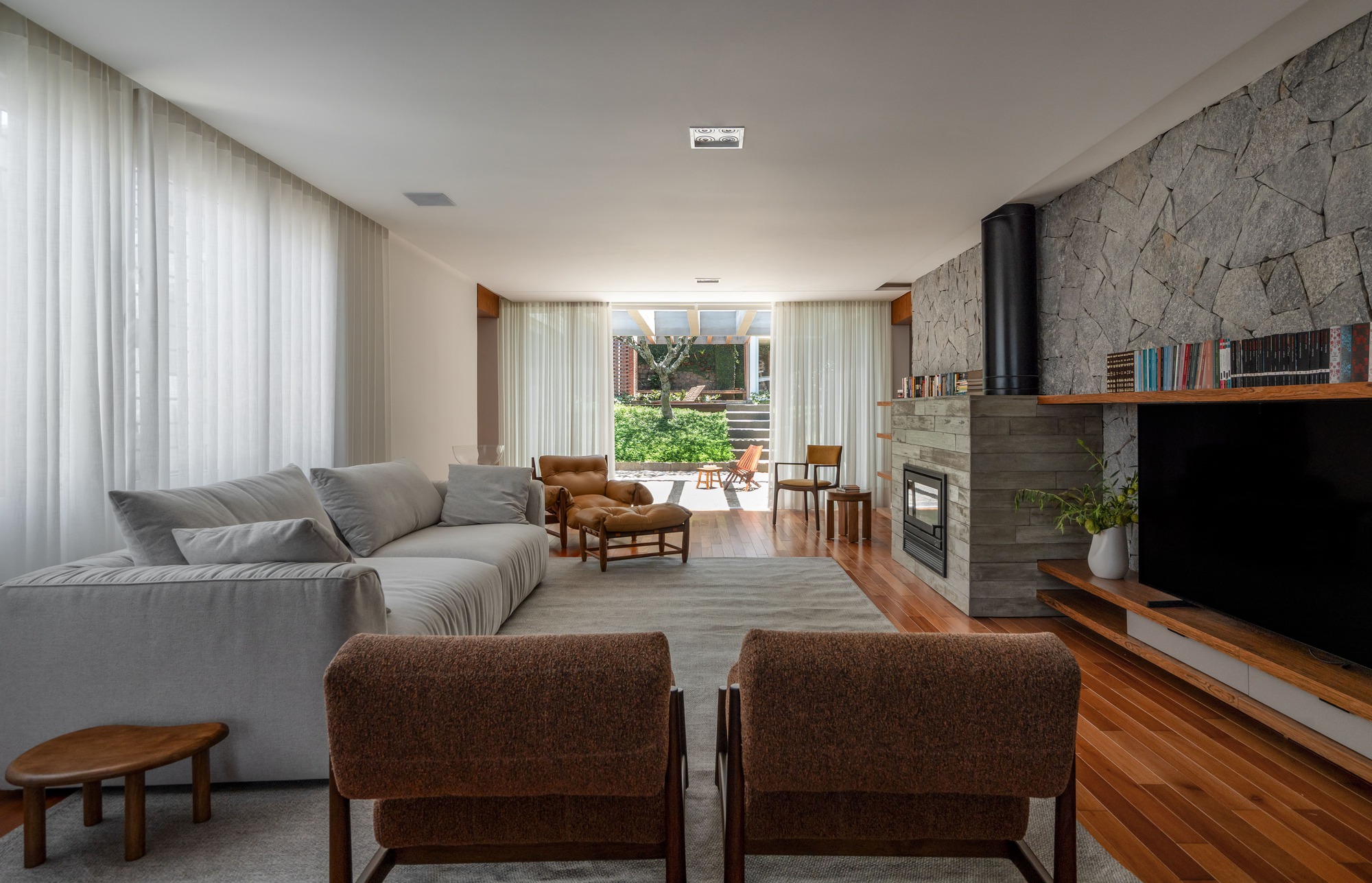 Living room with a pale sofa, brown armchairs and a stone fireplace wall, sheer curtains framing the garden beyond