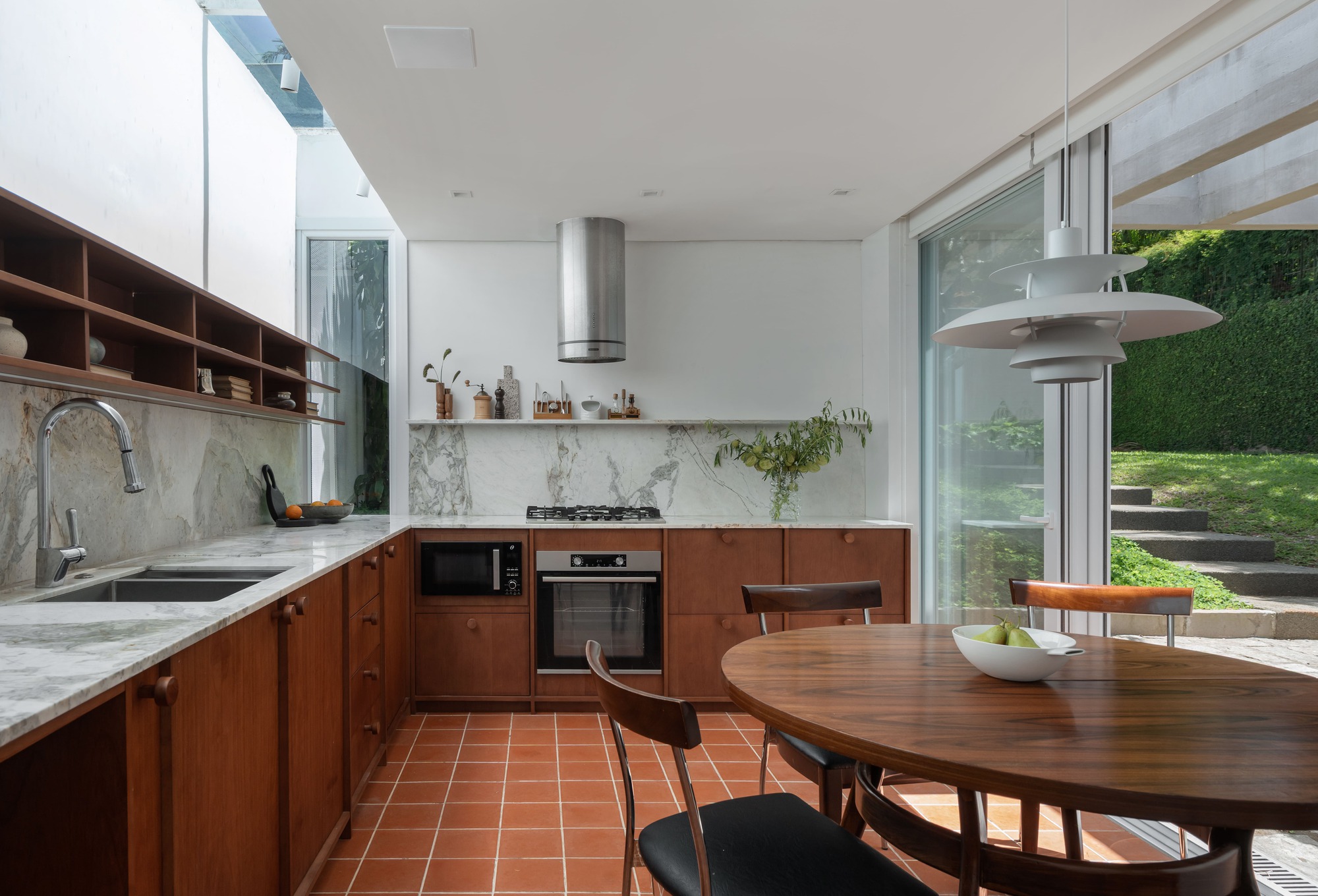 Kitchen with walnut cabinets, marble counter and terracotta floor tiles, a round wooden table beside glass doors opening to a green lawn
