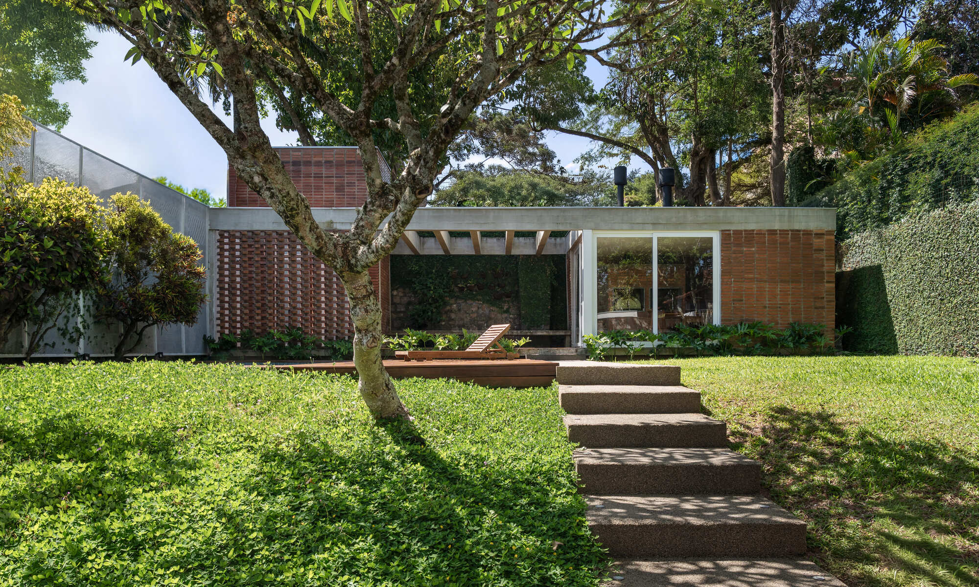 Garden facade of the brick and concrete pavilion seen across a lawn, with a stone stair and frangipani tree in dappled sun