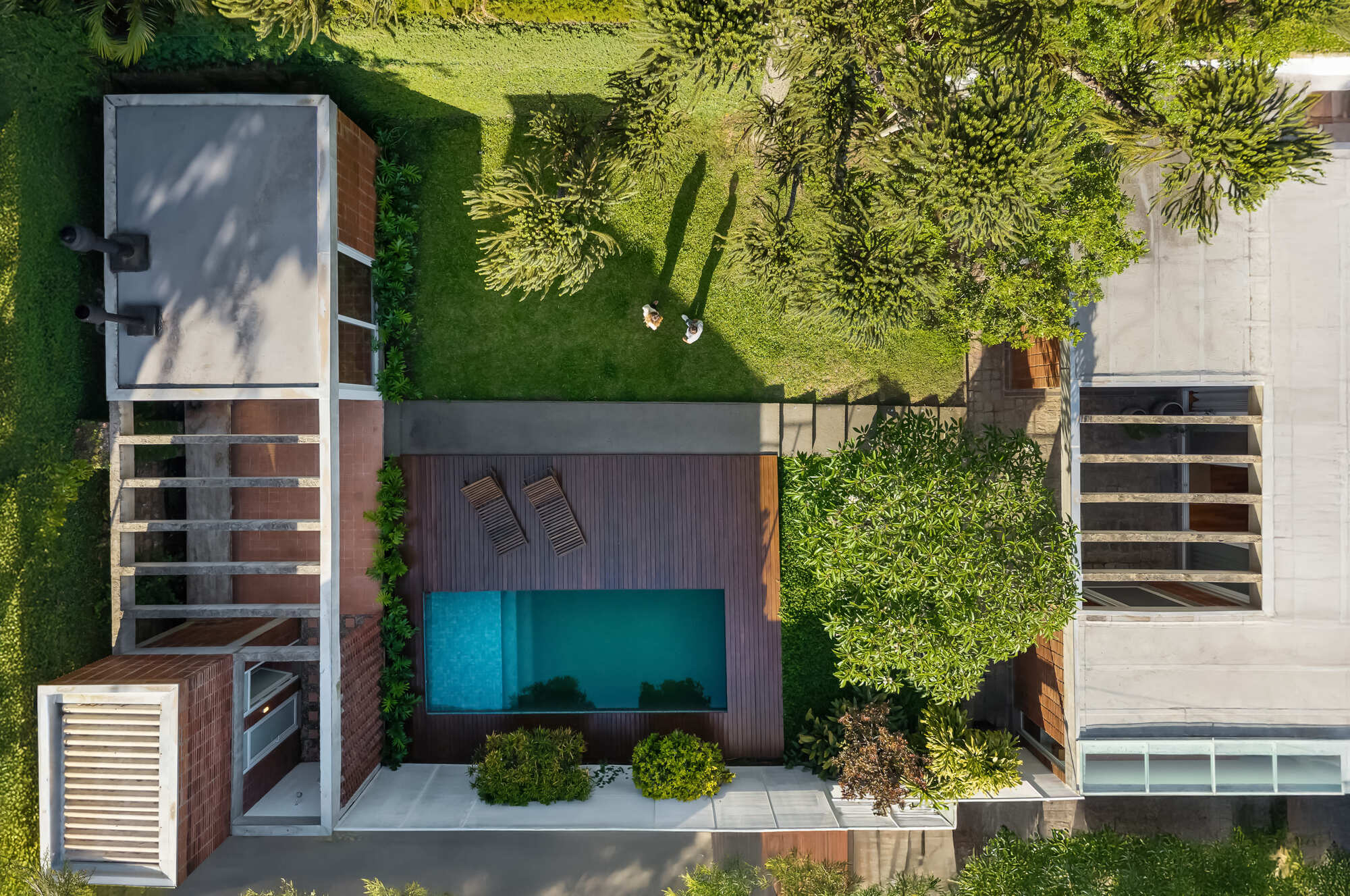 Top-down view of a timber deck and turquoise pool between brick-clad volumes and a flat roof, with tree shadows across the lawn