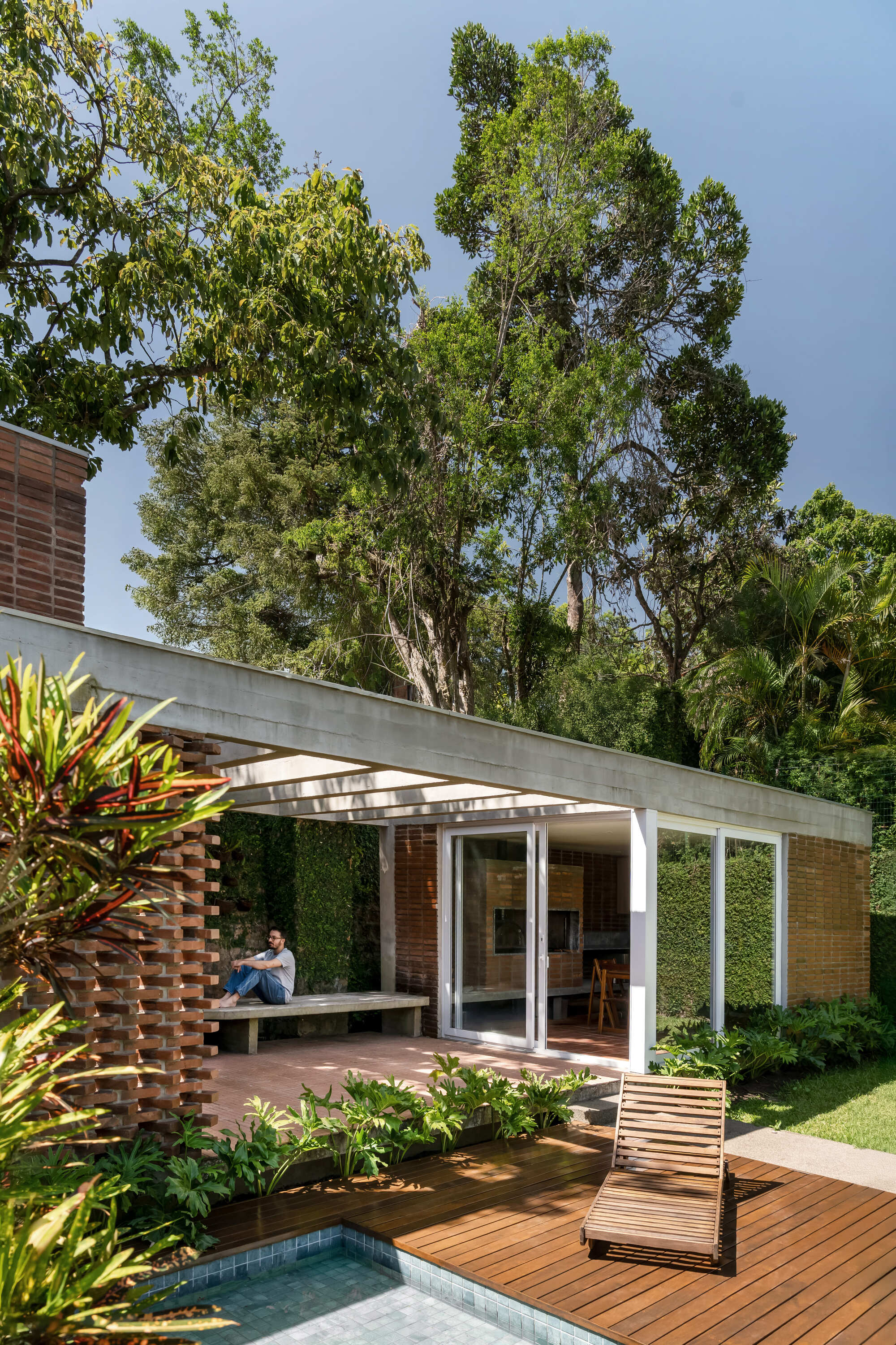 Covered brick porch with a concrete slab roof and a man seated on a bench, framed by tall trees above the pool deck