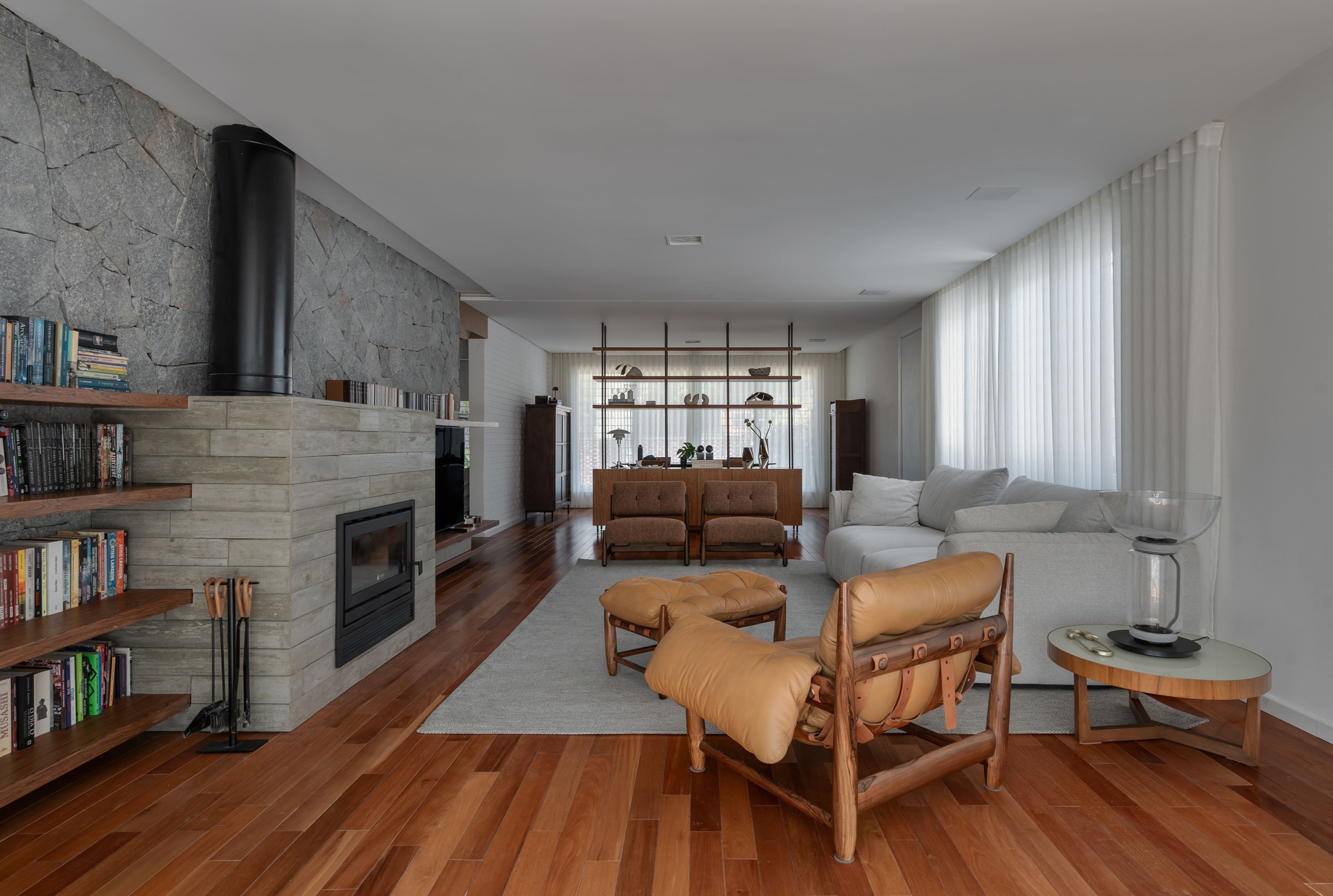 Living room with a stone chimney wall, built-in bookshelves, leather armchairs and sheer curtains over a warm hardwood floor