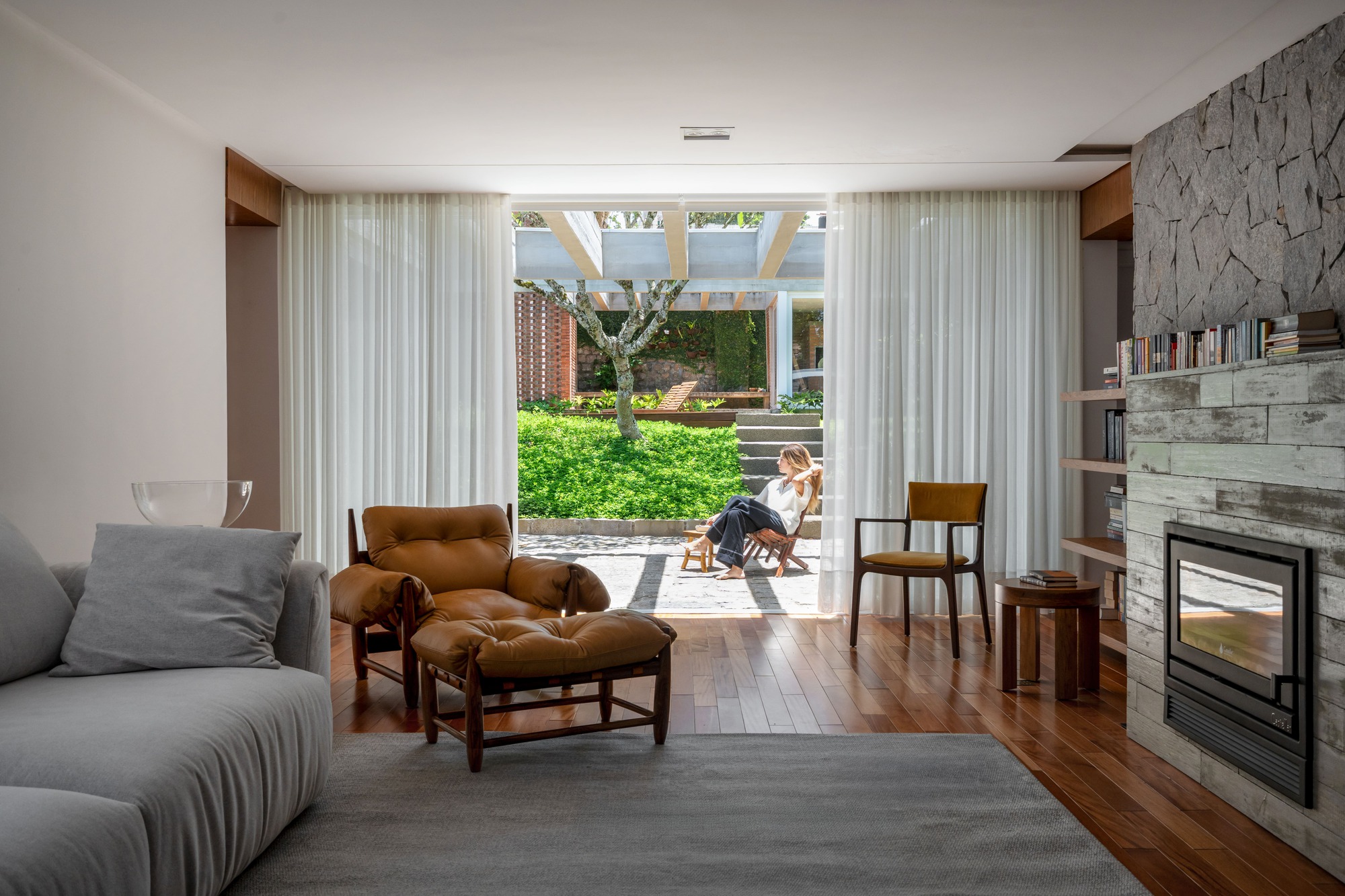 Living room seen through open sheer curtains toward a sunlit paved courtyard where a woman reclines in a folding chair