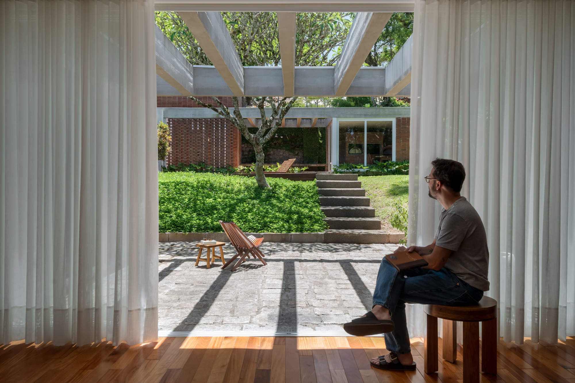 Man seated on a wooden stool between white curtains, looking across a paved courtyard to a concrete pergola and stepped lawn
