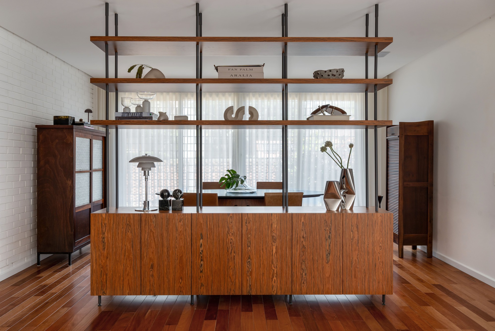 Freestanding wood sideboard topped by open steel-framed shelves, set against sheer curtains and a white brick wall