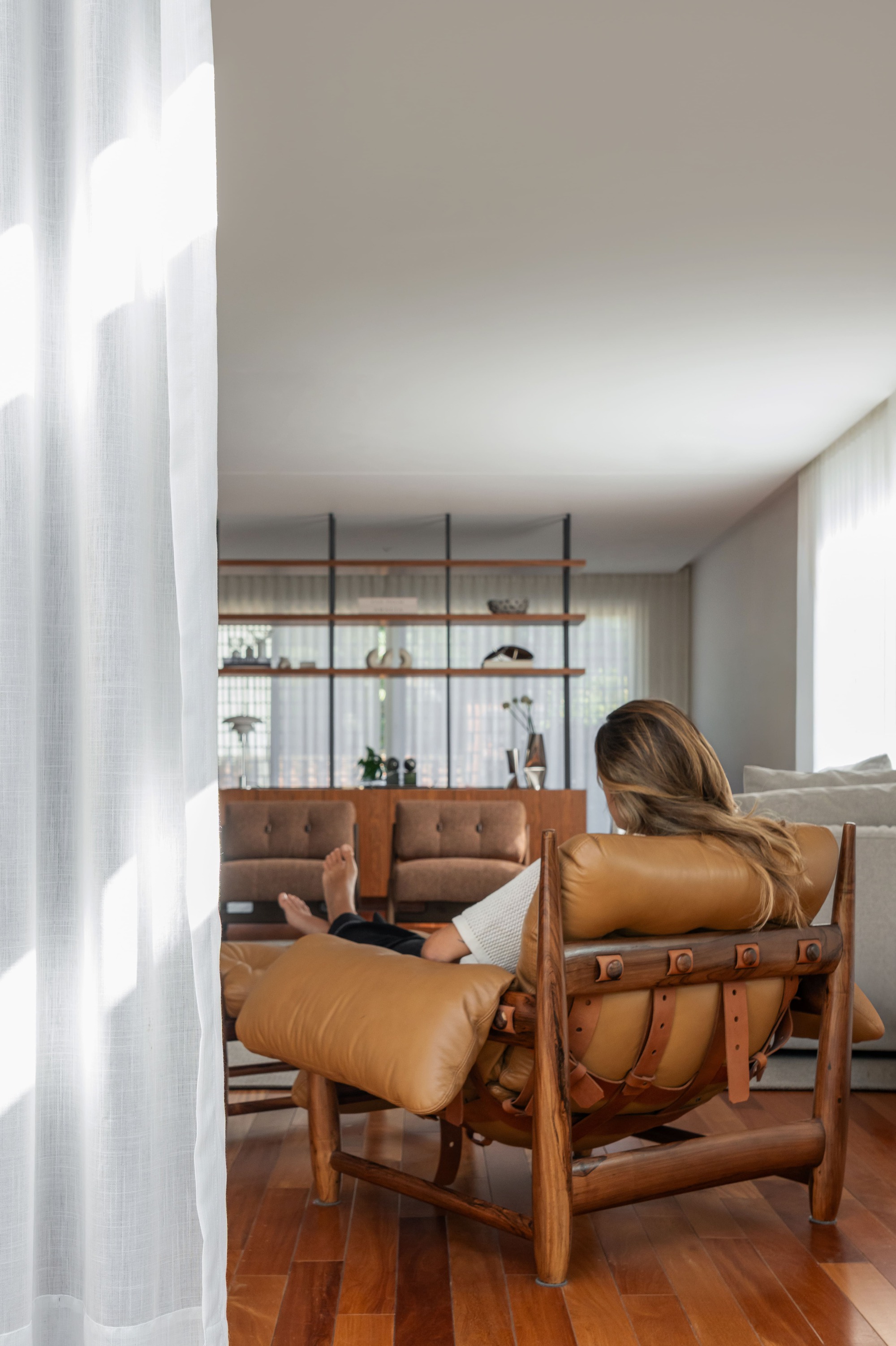 Woman reclining in a leather and timber lounge chair with ottoman, sheer curtain glowing beside her in daylight