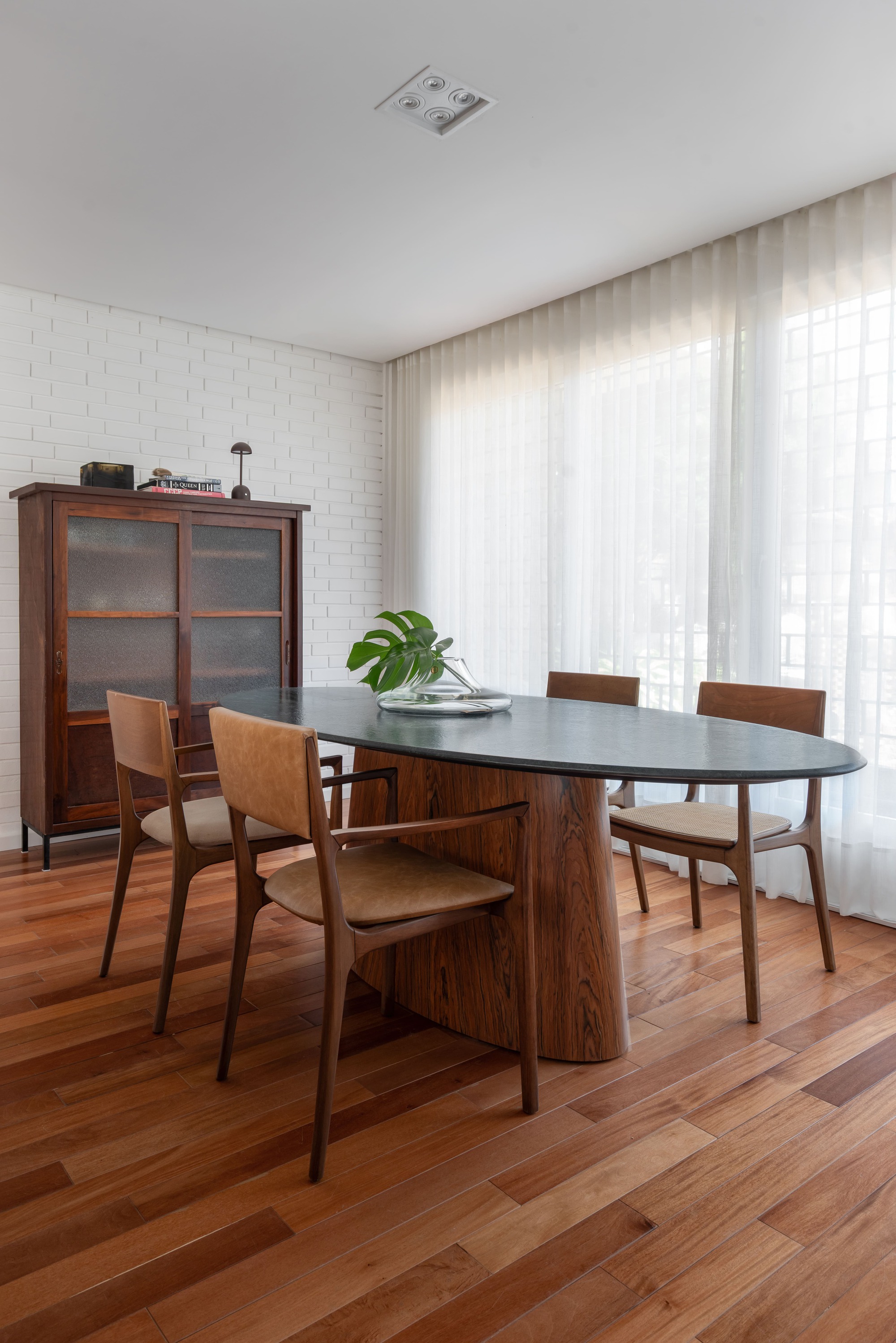 Oval dining table with a stone top and carved wood base, ringed by timber chairs beside sheer curtains